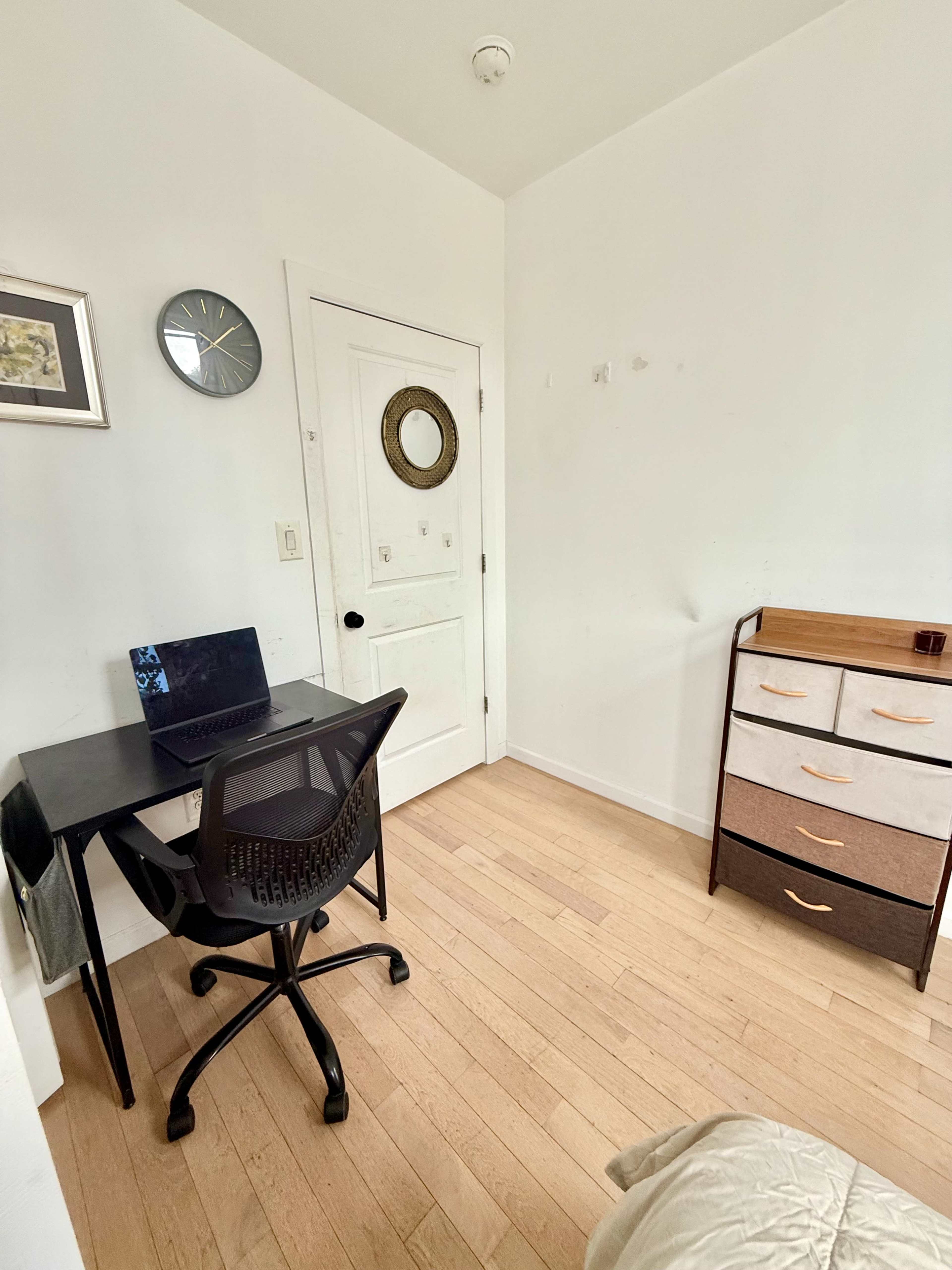 The image shows a small, sparsely furnished room with a desk and chair, a dresser, and a closed door.