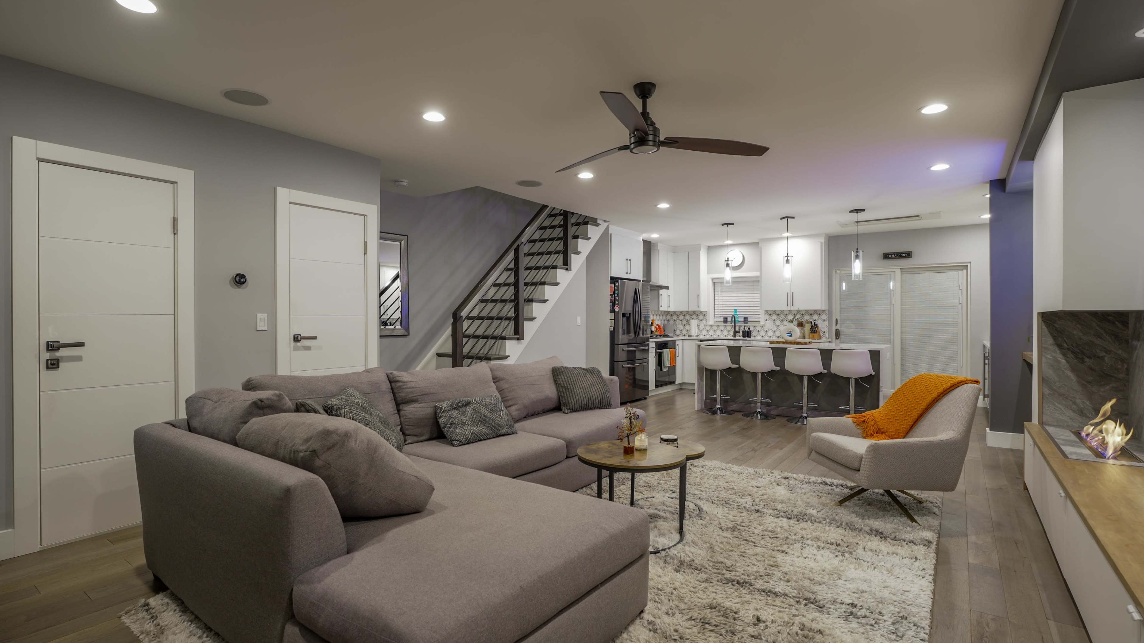 The image shows a modern living room with a sectional sofa, a small coffee table, and an adjoining kitchen area, featuring a staircase in the background.