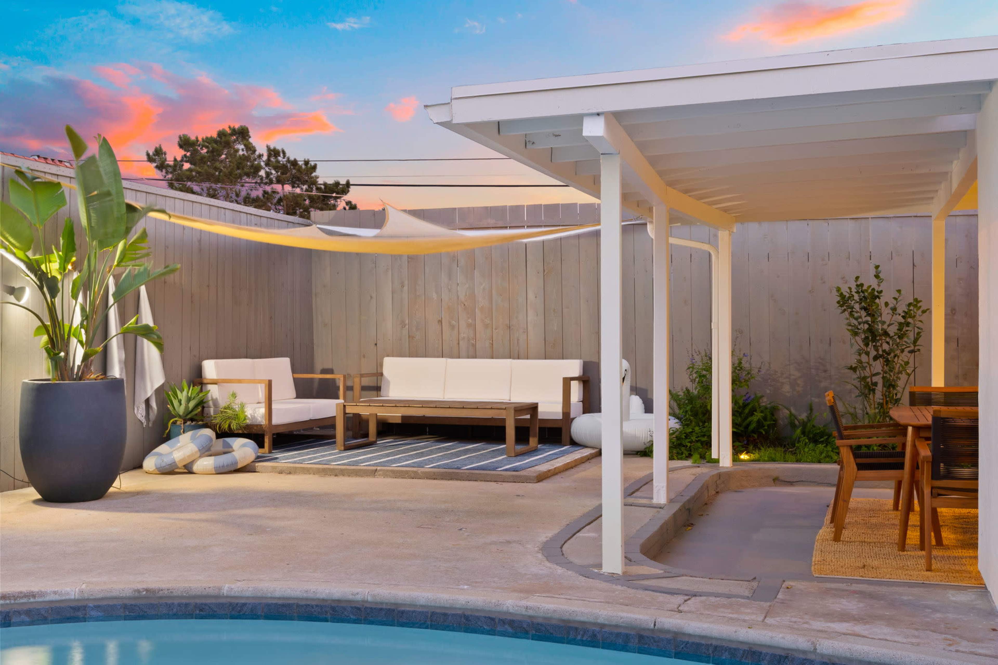 The scene features a modern outdoor living area with a pool, comfortable seating, and a dining space, surrounded by wooden fencing and plants under a colorful sunset sky.