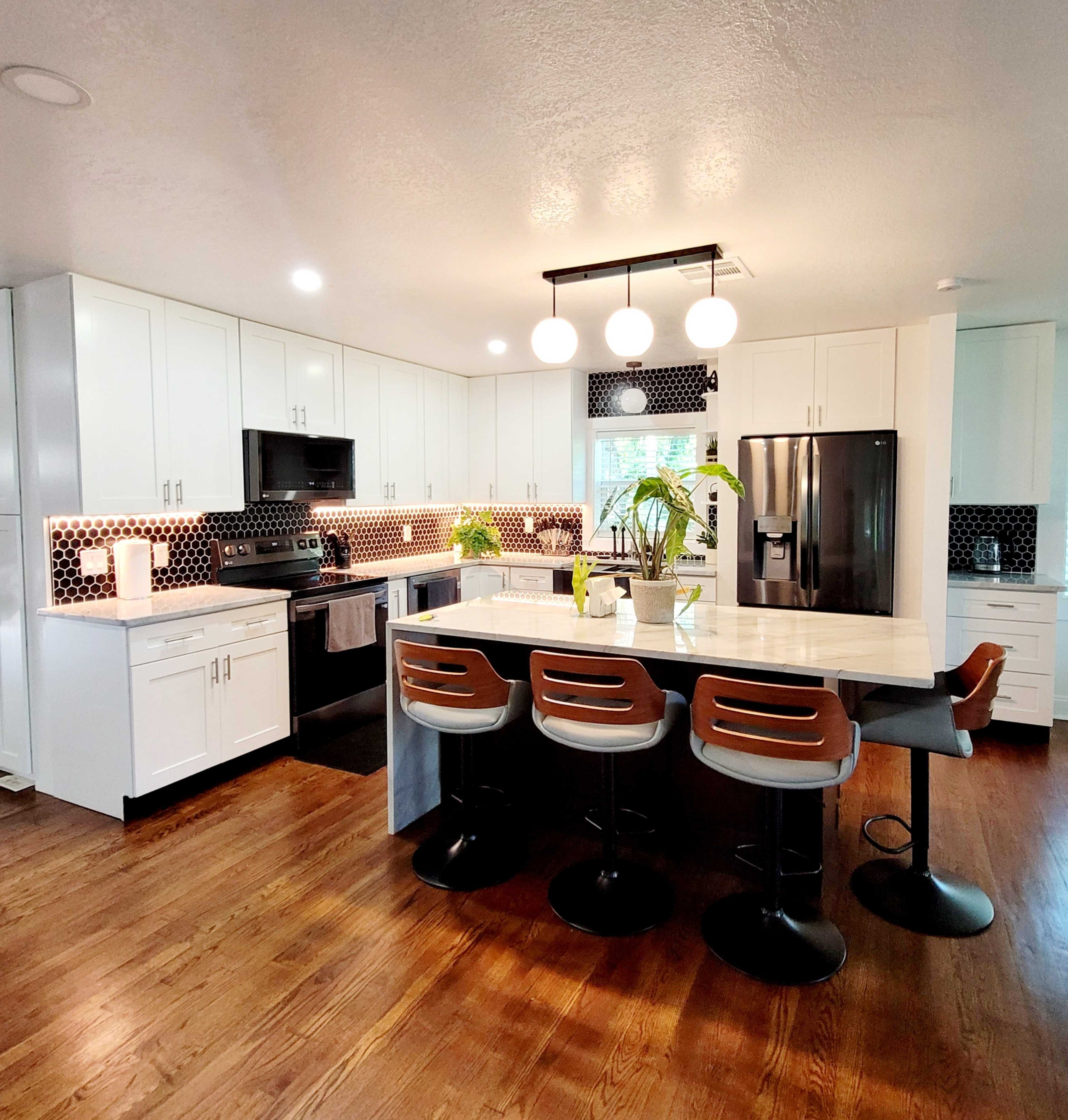 The image shows a modern kitchen with white cabinets, a marble island with bar stools, and a mix of dark hexagonal tiles and stainless steel appliances.