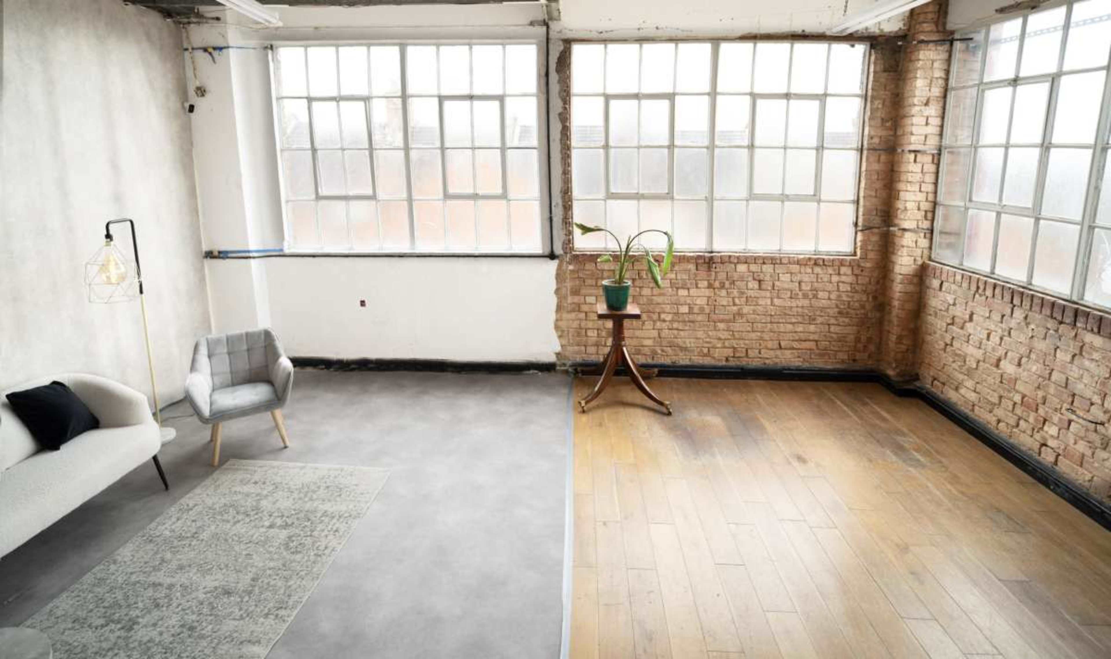 The image shows a spacious room divided into two sections, one with a light grey floor and a sofa, and the other with wooden flooring and a potted plant on a small table.