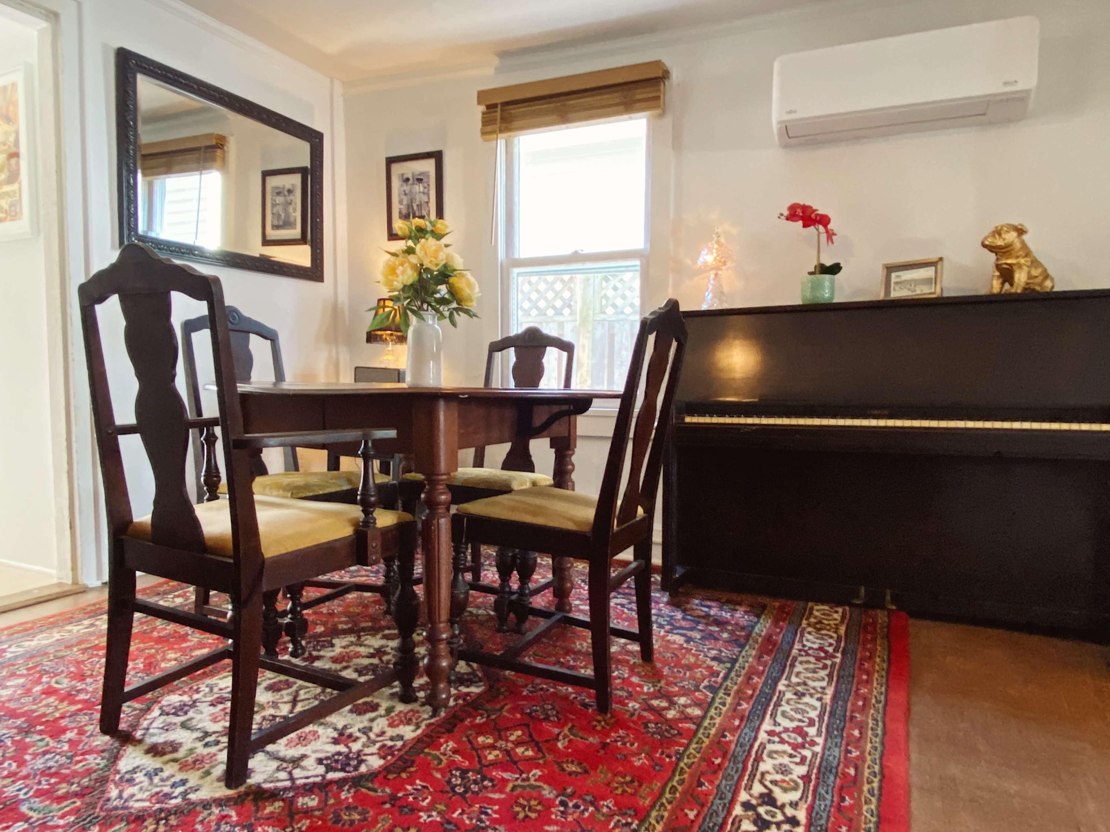 A dining area features a wooden table with four chairs, a piano, and a rug with intricate patterns.