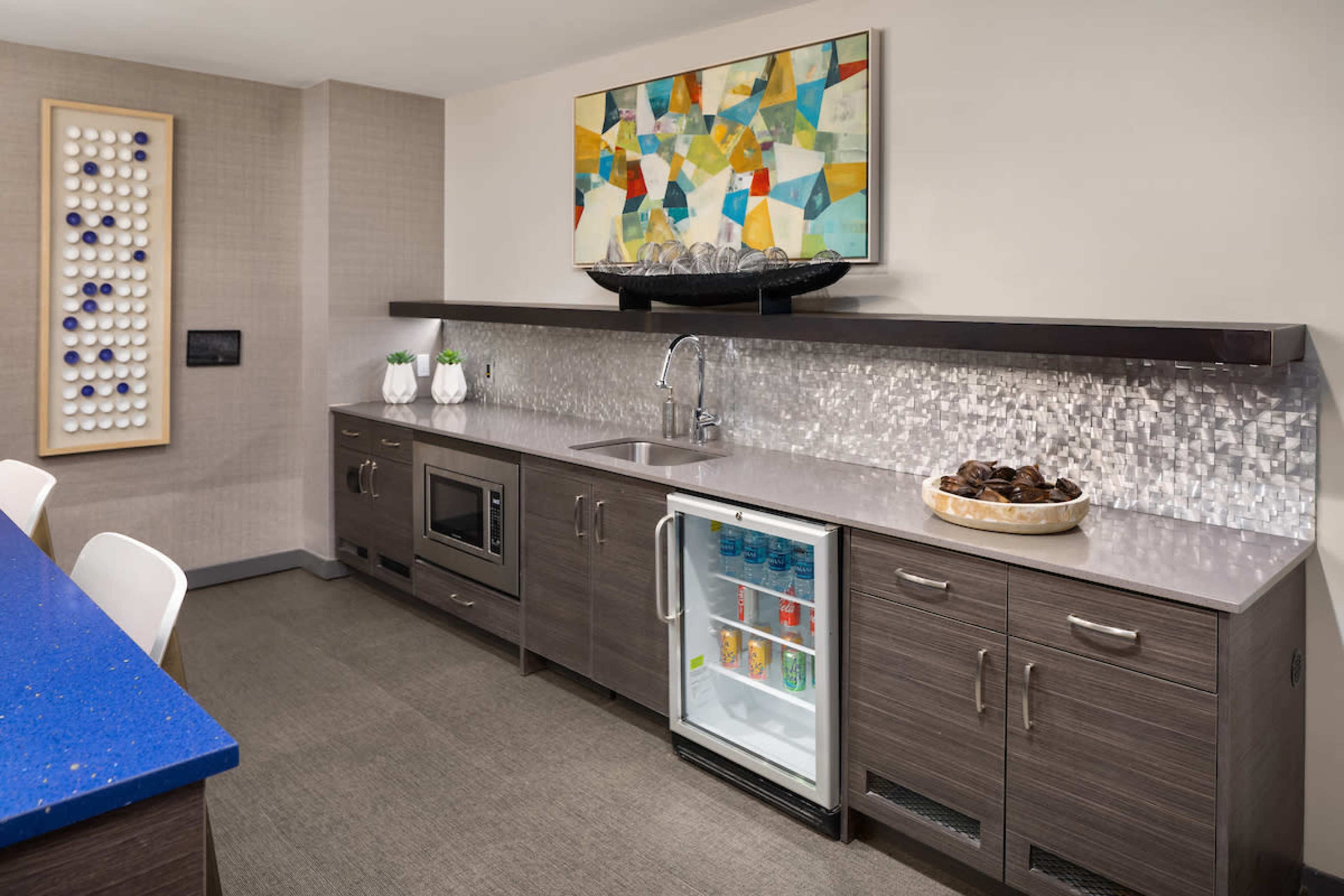 The image shows a modern kitchen with a sleek countertop, a small refrigerator, a microwave, and decorative elements above the sink.