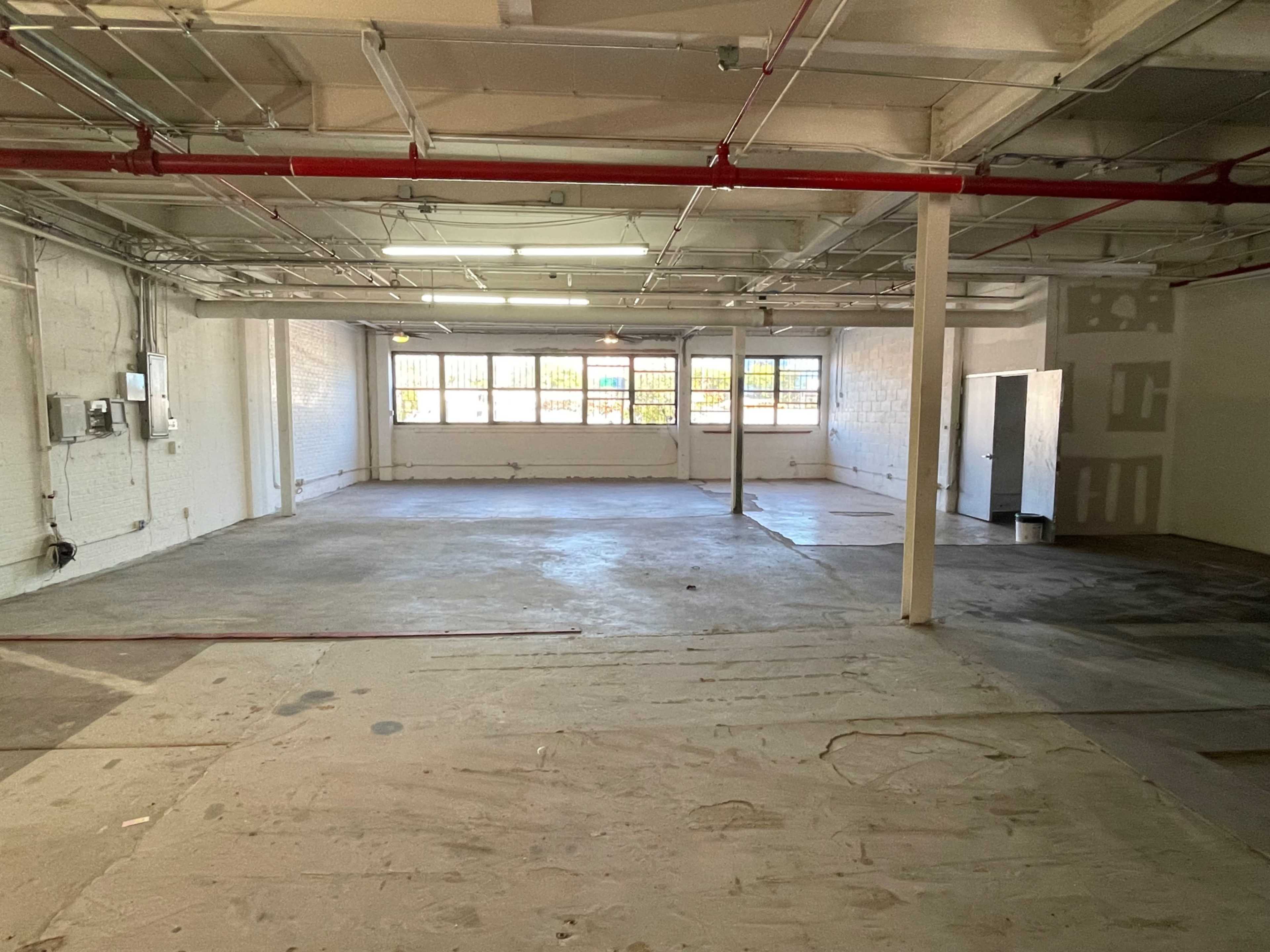 The image shows a spacious, empty room with concrete floors, exposed pipes, and large windows allowing natural light.