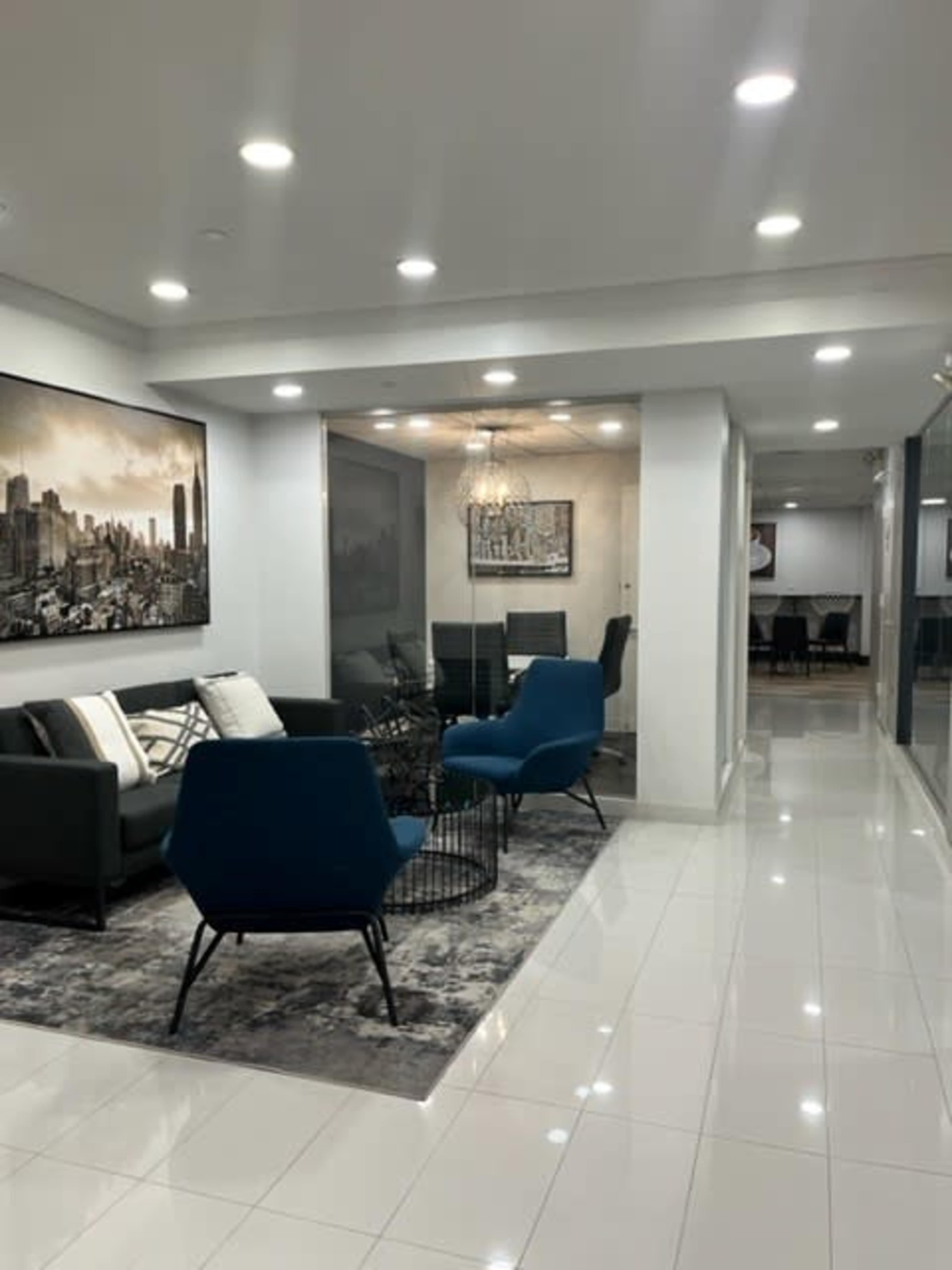 The image shows a modern interior space featuring a seating area with blue chairs and a large wall art piece, leading to a dining area visible in the background.