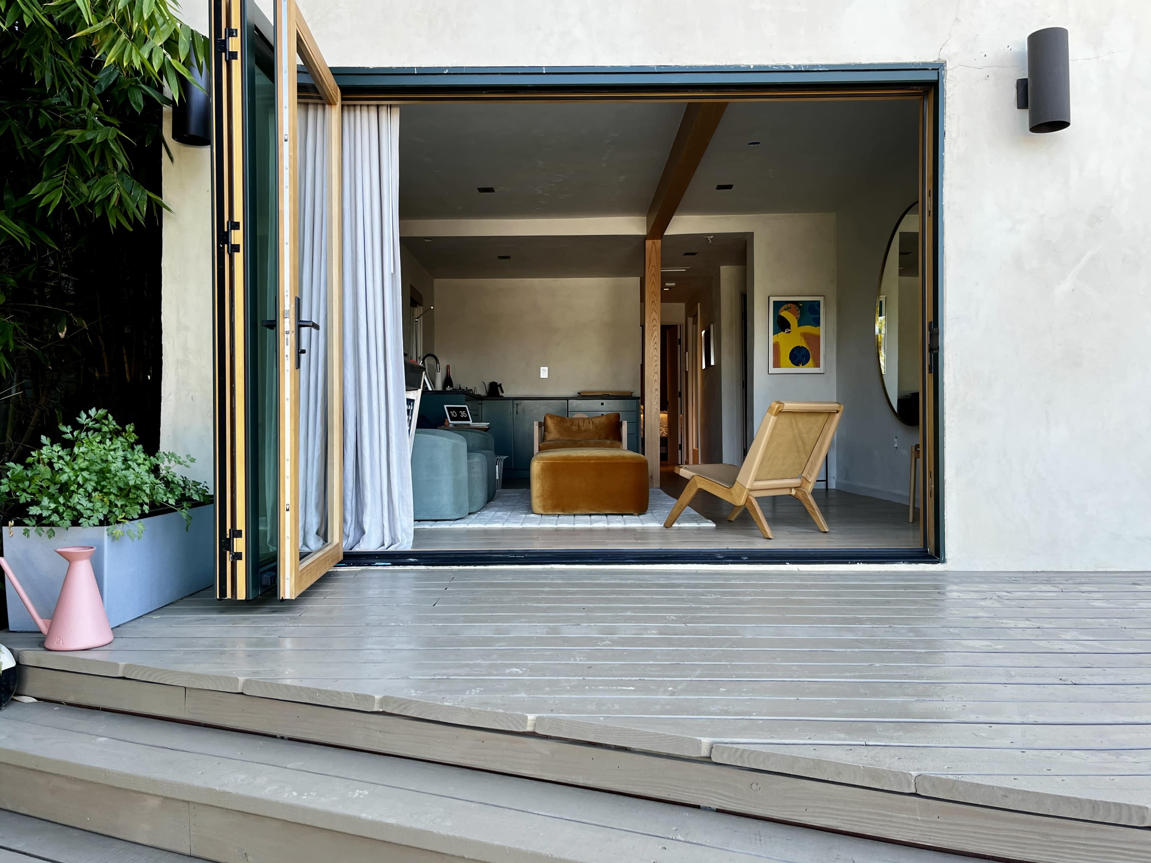The image shows a wooden deck with a sliding glass door leading into a modern living room that features a brown sofa and a light-colored rug.