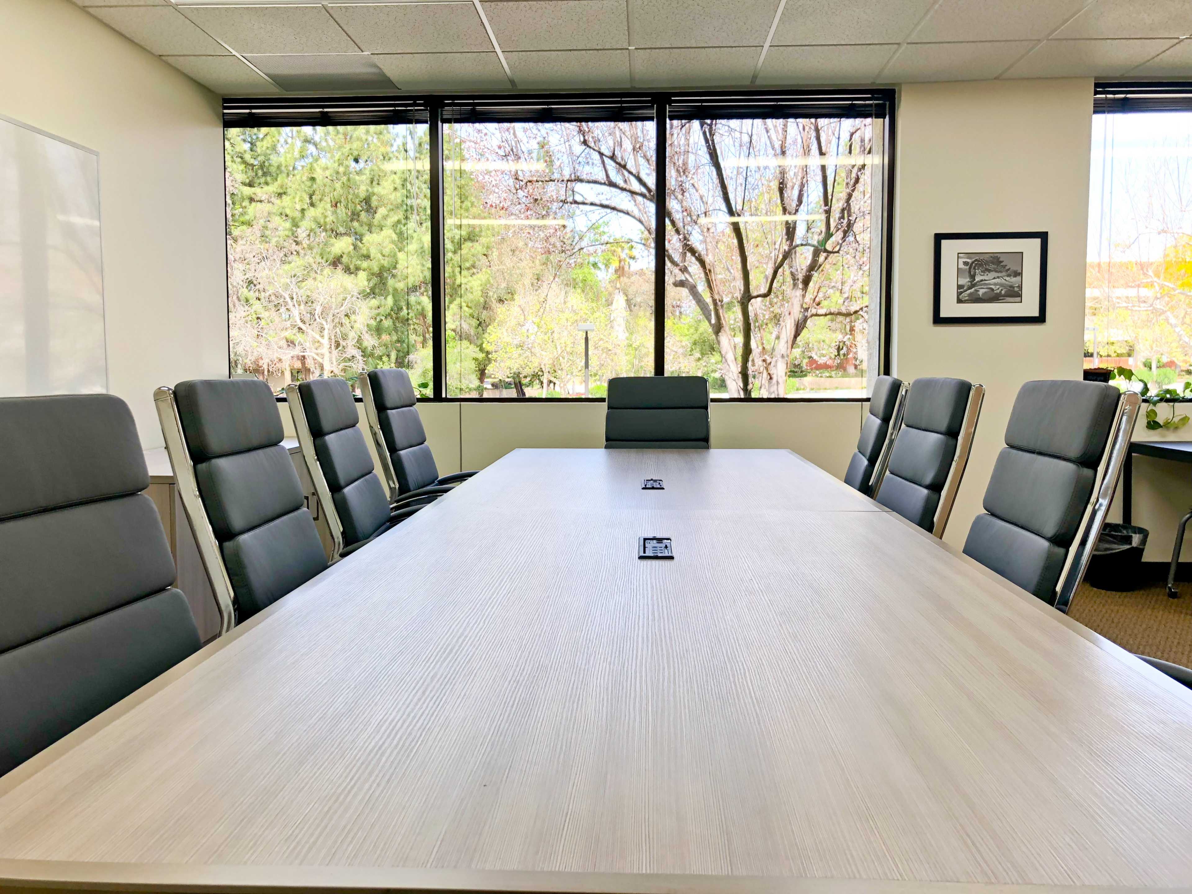 A modern conference room with a large wooden table surrounded by gray chairs and a backdrop of large windows showing greenery outside.