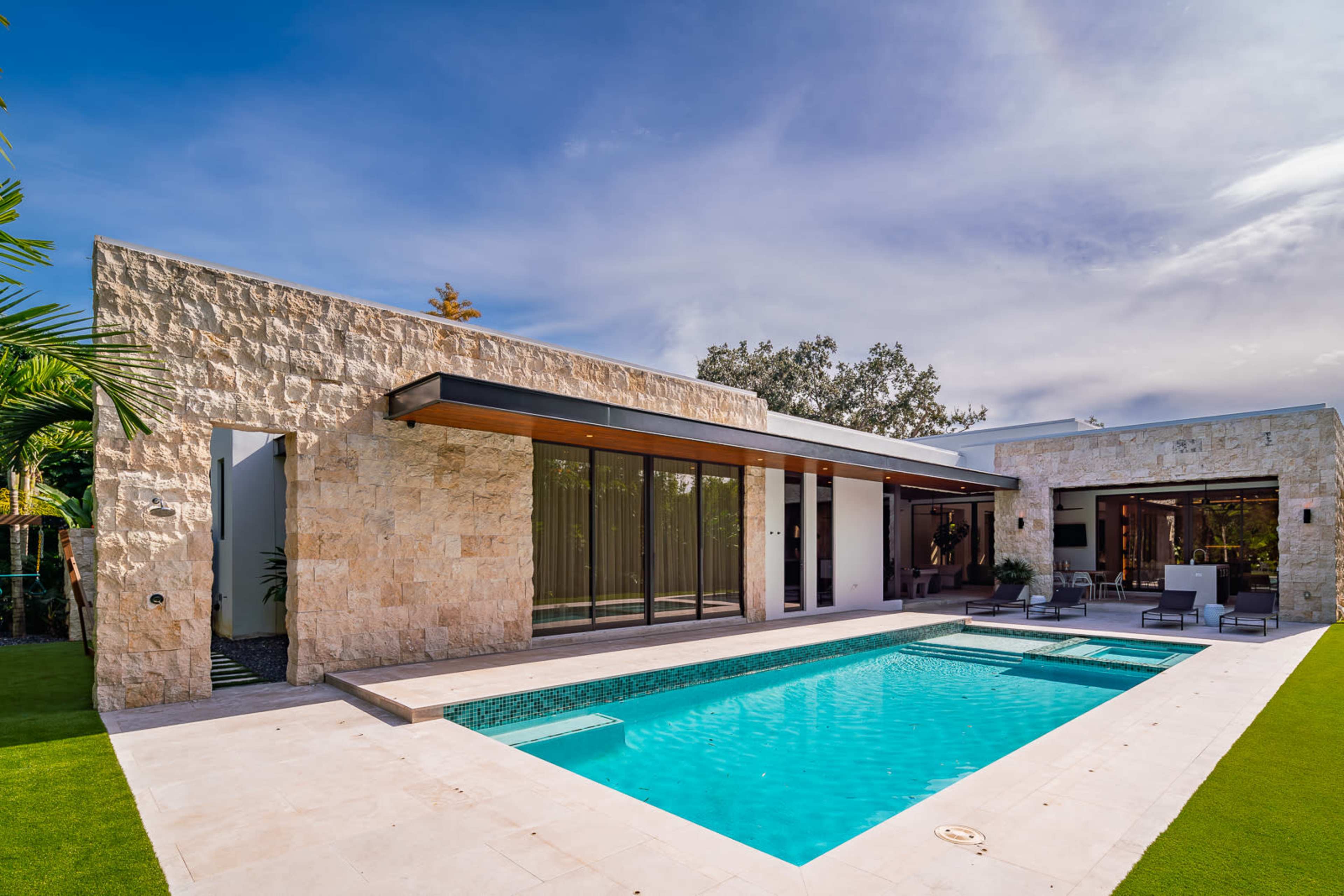 The image shows a modern house with a stone facade, large glass doors opening to a swimming pool, and a landscaped lawn.
