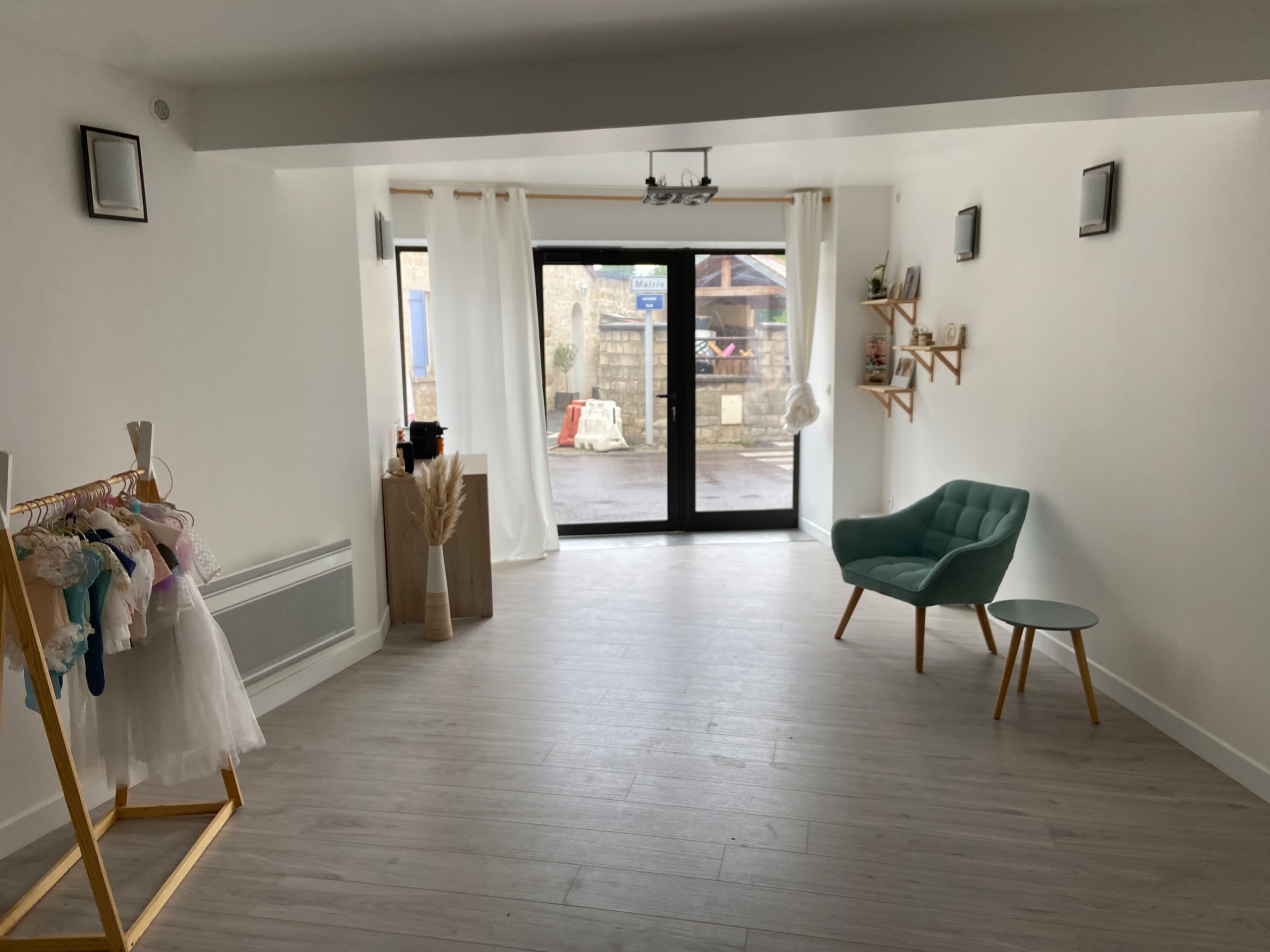 The interior of a minimalist shop features a green chair and small table, a clothing rack with garments, and large windows facing a street.