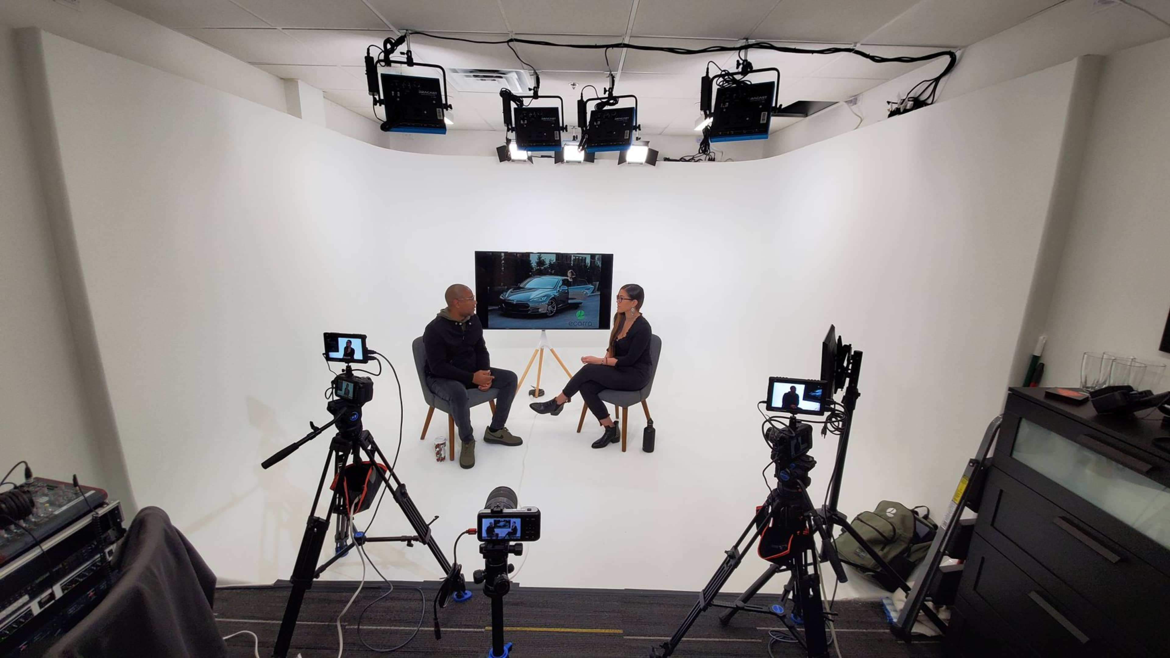 A man and a woman sit across from each other in a studio setting with multiple cameras and bright lights, while a large screen displays an image of a car.