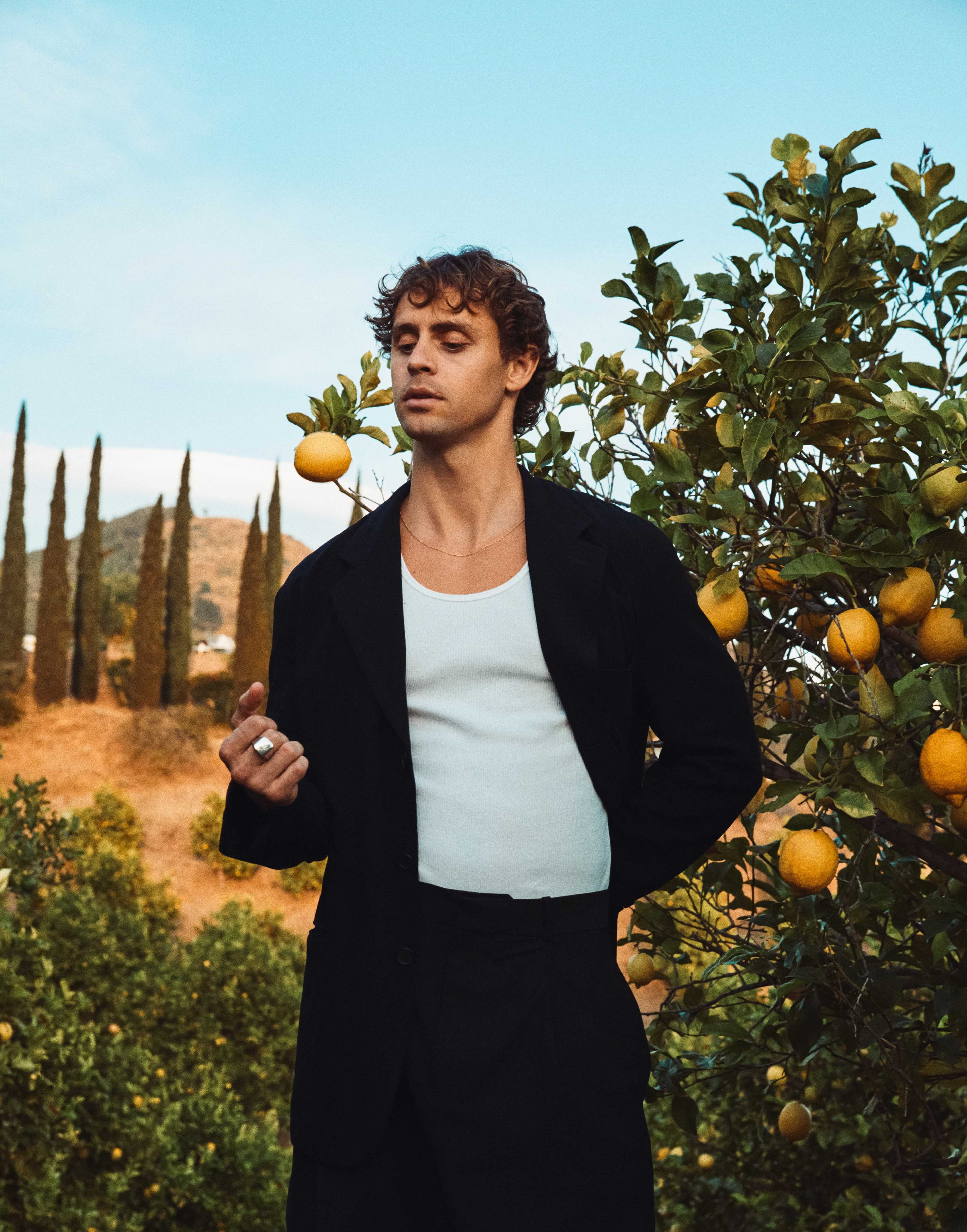 A person in a black blazer stands among lemon trees, with luscious yellow lemons and tall cypress trees in the background against a blue sky.