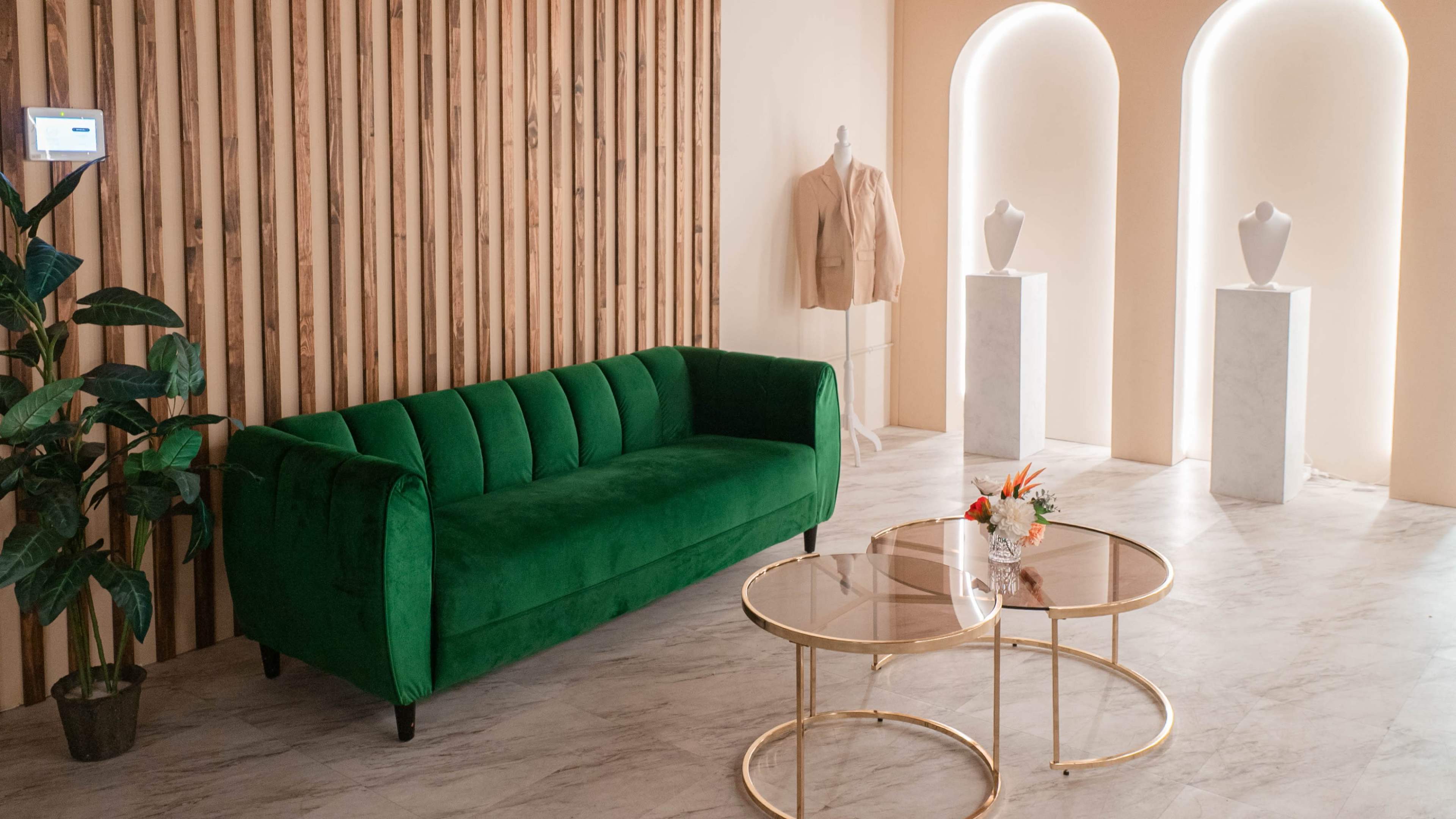 The image shows a modern interior space featuring a green velvet sofa, a gold and glass coffee table, and two white display pedestals against a backdrop of wooden slat walls.