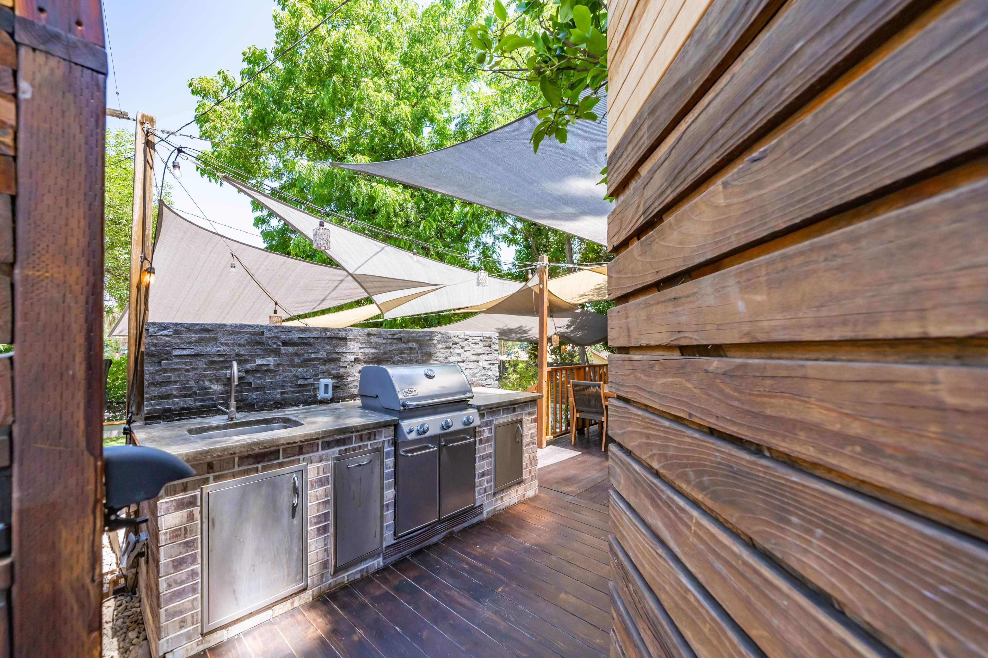 The image shows an outdoor kitchen area with a stainless steel grill, stone countertops, and shaded by fabric canopies.
