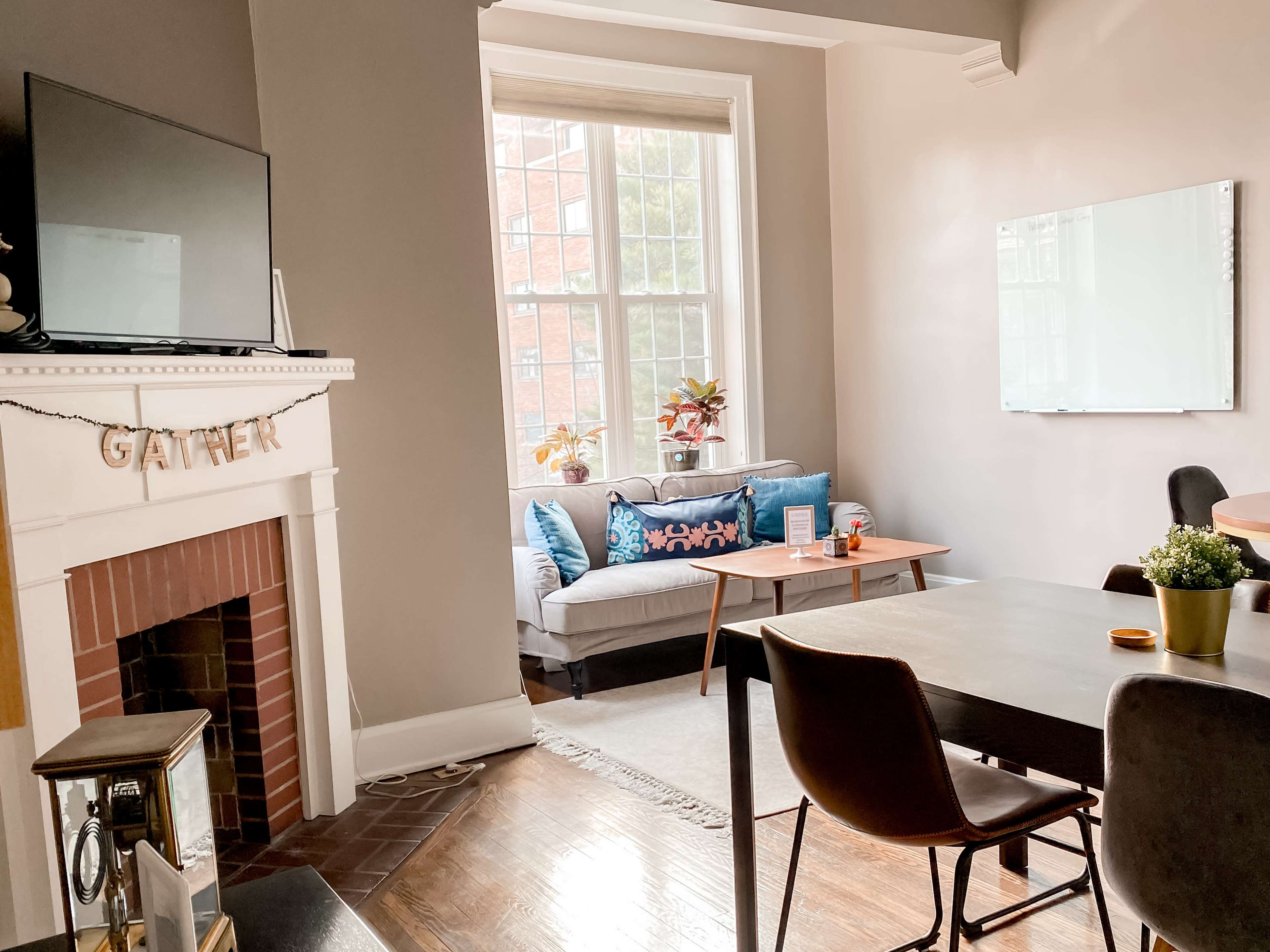 The image shows a cozy living space featuring a fireplace with a "GATHER" banner, a sectional sofa near a large window, a coffee table, and a dining table with chairs.