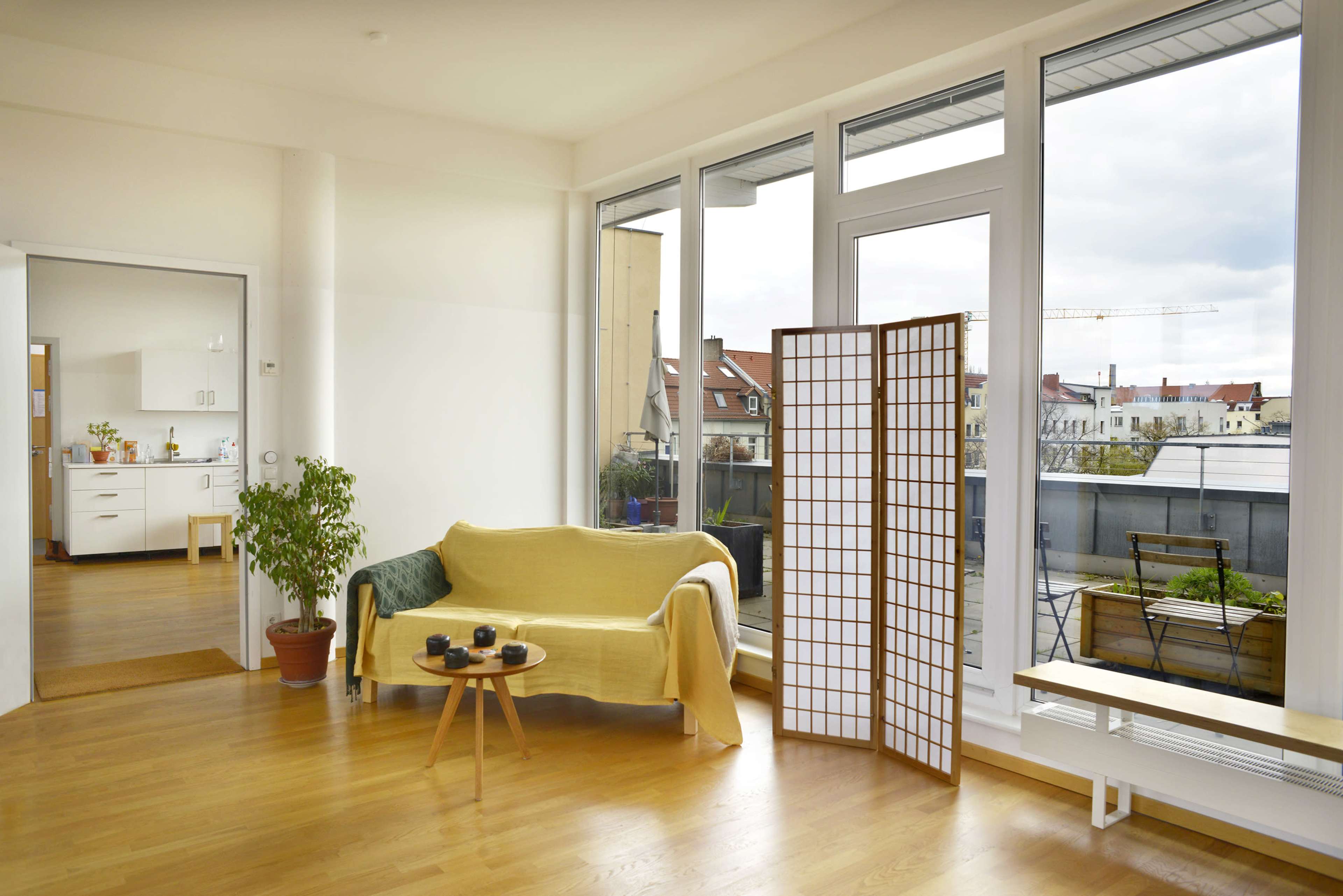 The image shows a brightly lit living room with a yellow couch, a wooden table with tea cups, and a room divider by a large window overlooking a cityscape.