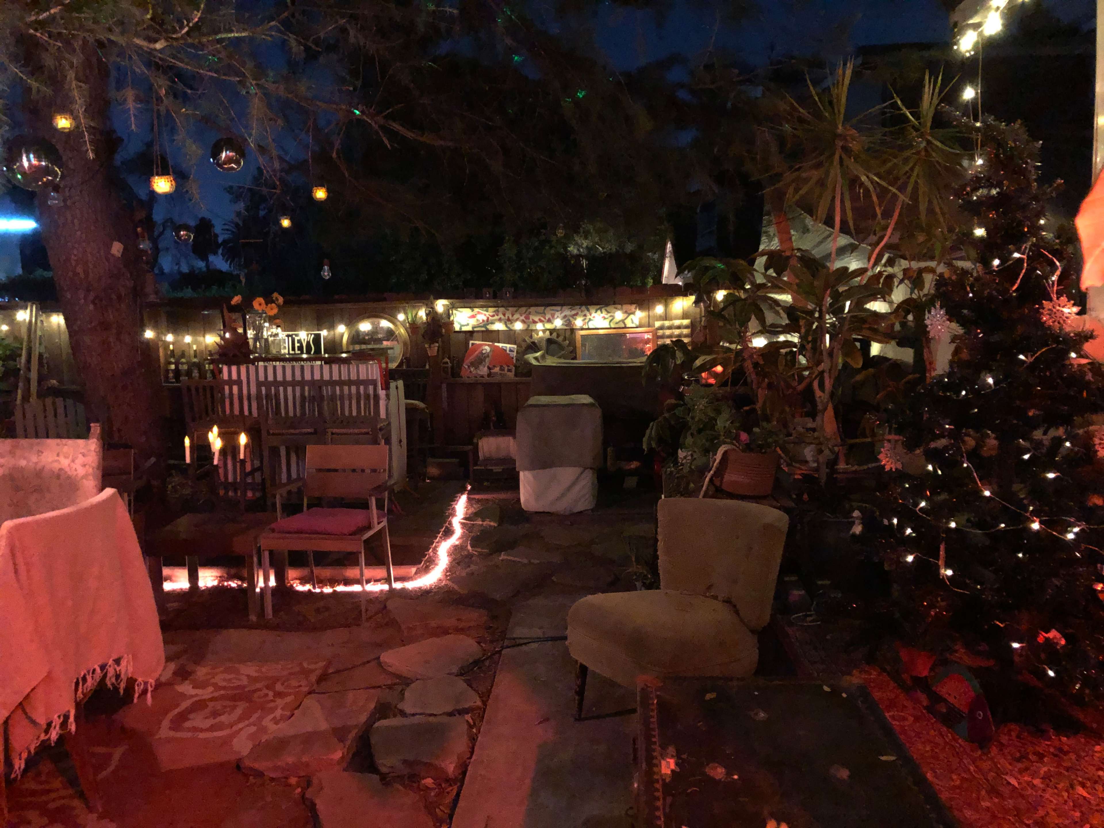 The scene depicts a dimly lit outdoor space with seating arrangements, decorative lights, and a small Christmas tree.