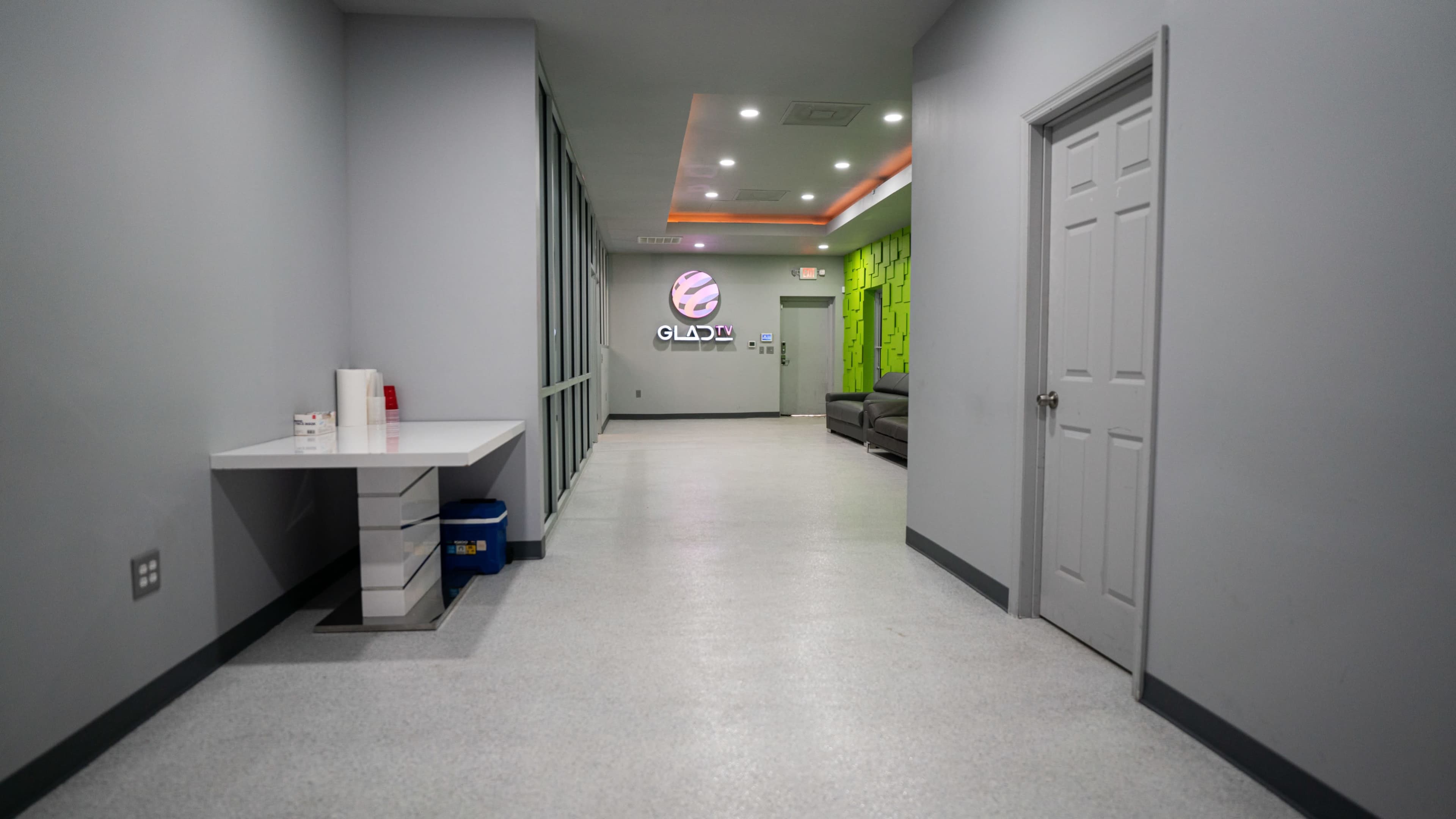 A modern hallway with grey walls and a reception area featuring a logo, leading to a green accent wall and a gray sofa.