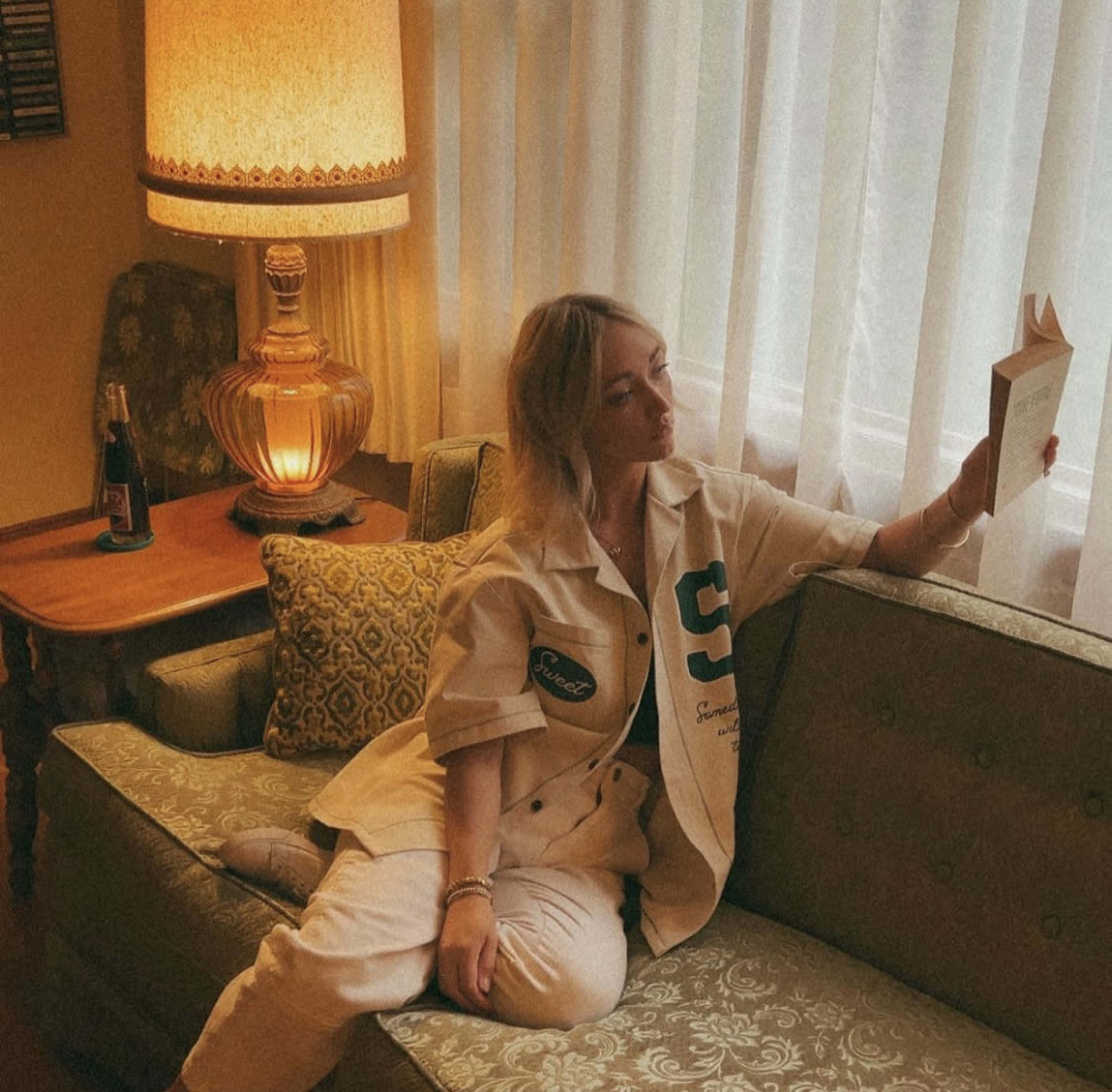 A woman sits on a vintage couch, reading a book next to an illuminated lamp and a bottle on a side table.
