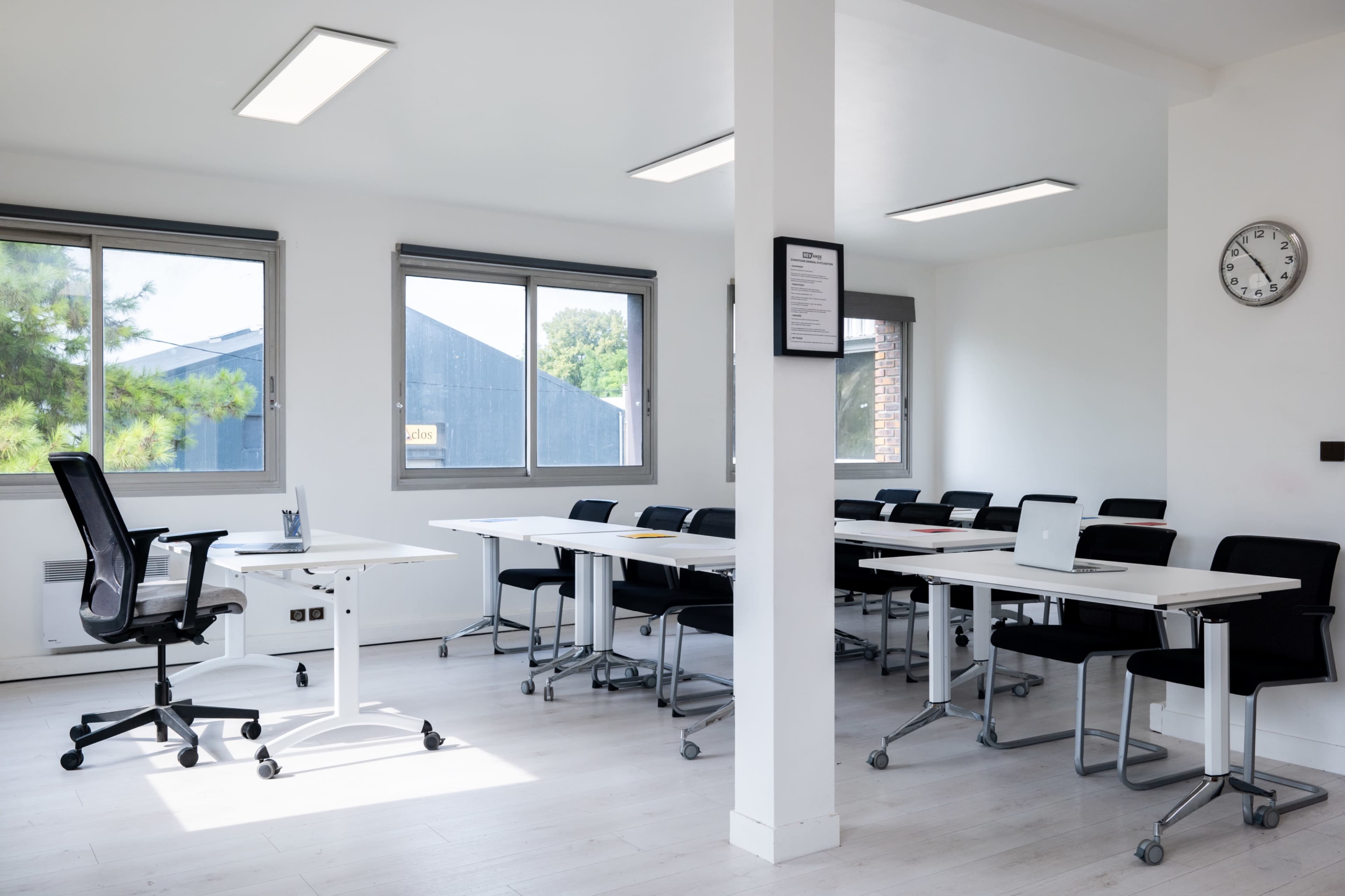 A bright, modern meeting room with a row of tables, black chairs, and large windows.