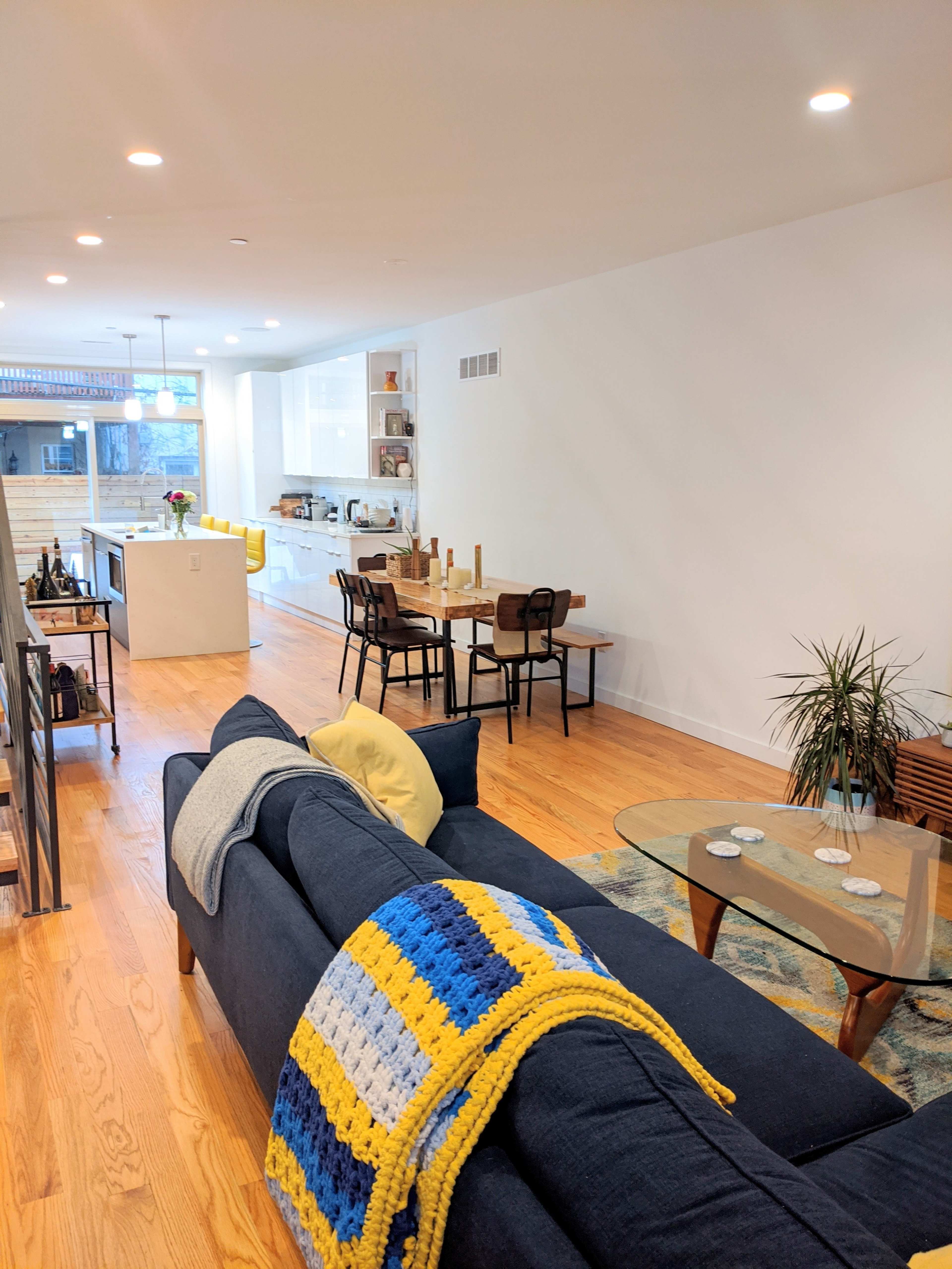 A modern living area features a blue sofa with a yellow and blue blanket, a glass coffee table, and a dining area with a wooden table set for meals.