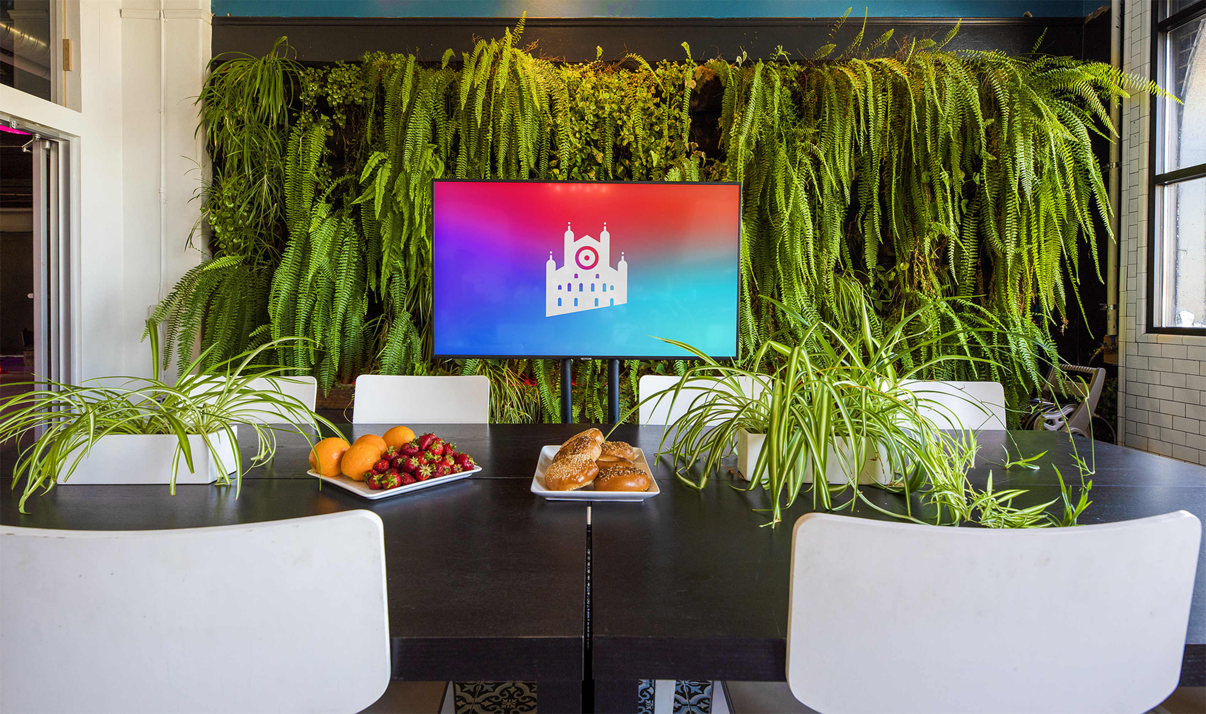 A dining table is set with fruit and pastries in front of a large screen displaying a colorful logo, surrounded by lush greenery on the walls.