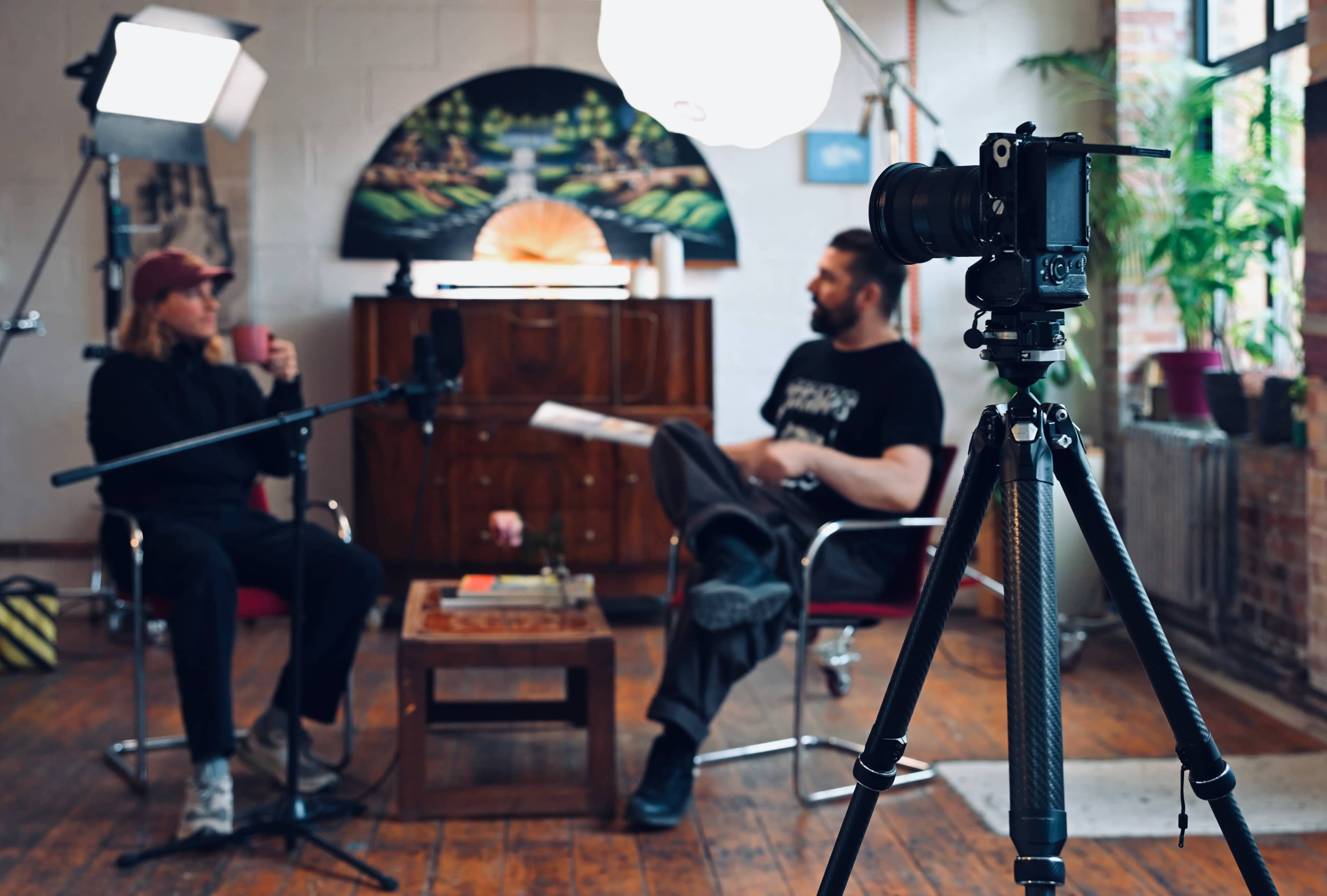 A camera on a tripod captures a conversation between two people in a well-lit, rustic studio setting with plants and artwork in the background.