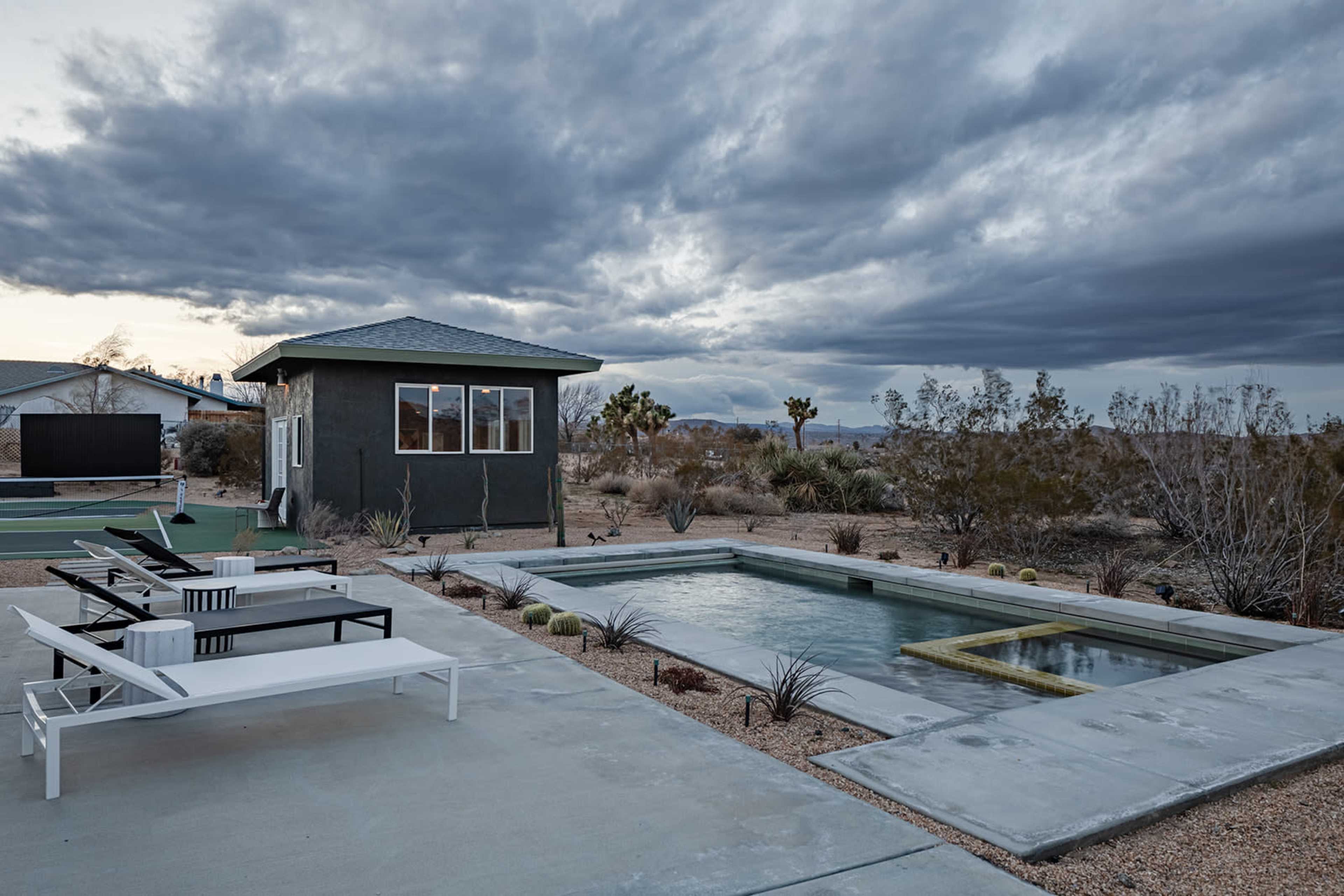 A swimming pool surrounded by desert landscaping and lounge chairs is situated next to a small building under a cloudy sky.