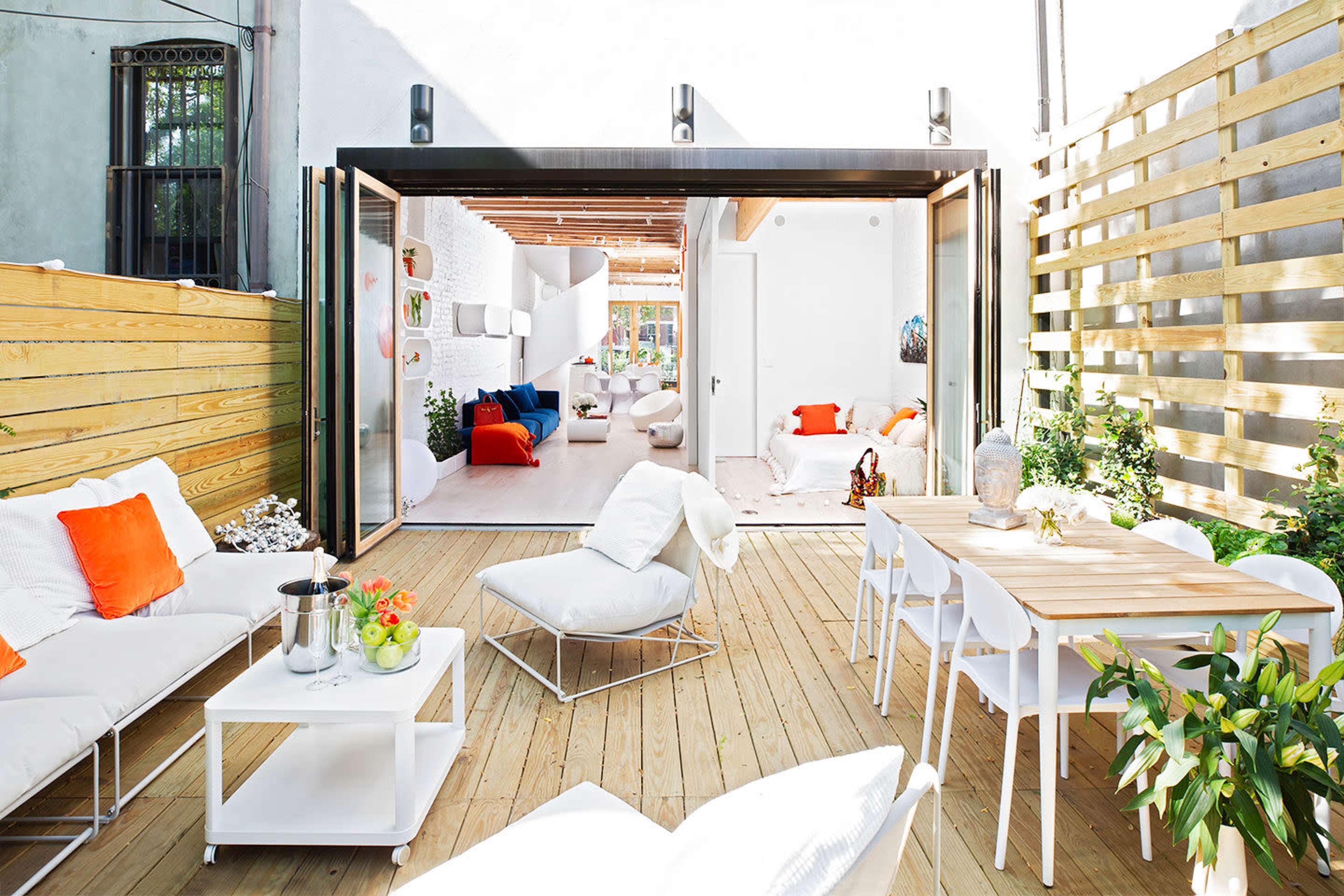 A modern patio area with white seating and a wooden dining table, leading to an open-space interior with large glass doors.