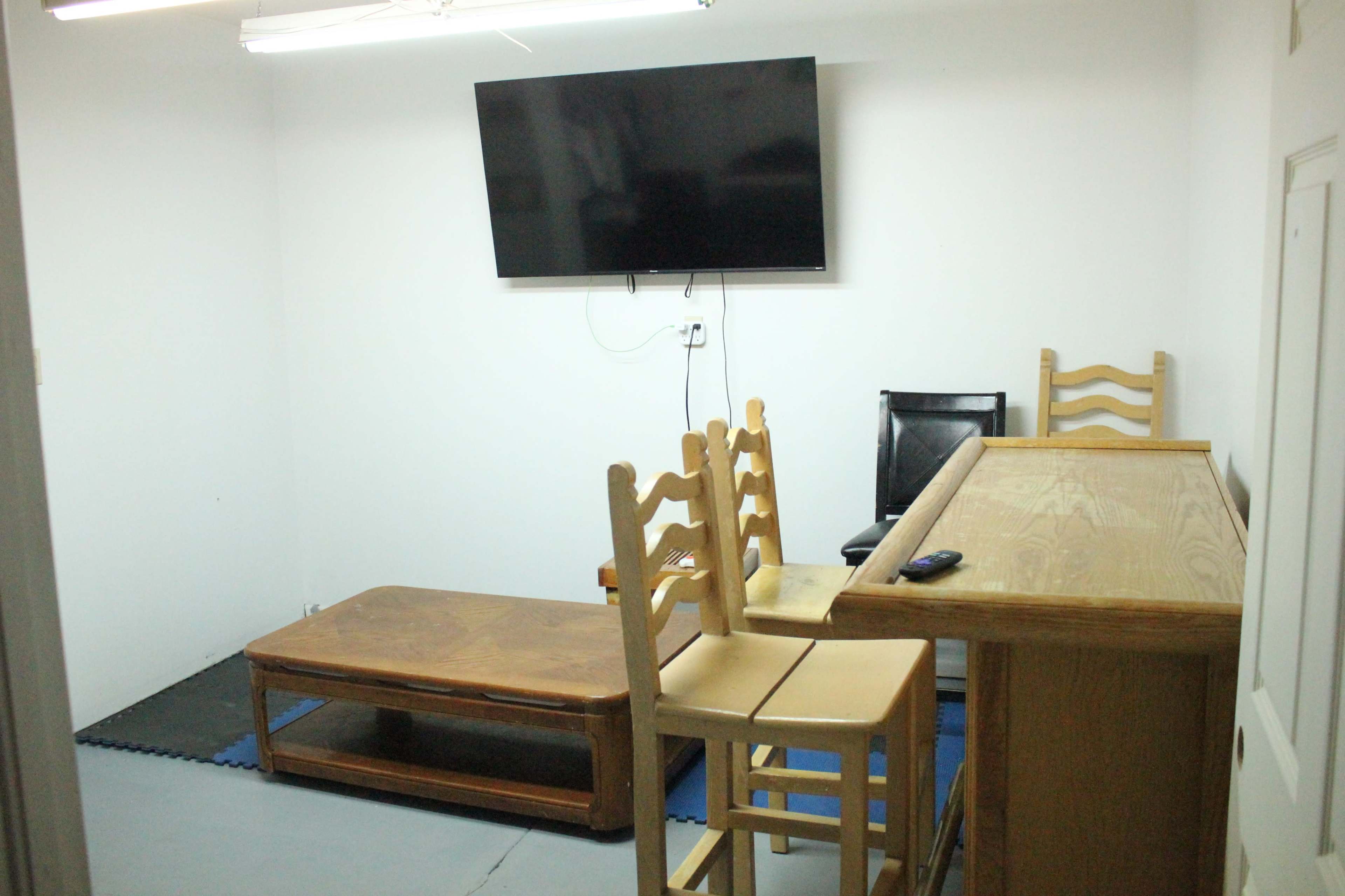 The image shows a small room with a wall-mounted TV, a wooden bar, a few chairs, and a low table on the floor.
