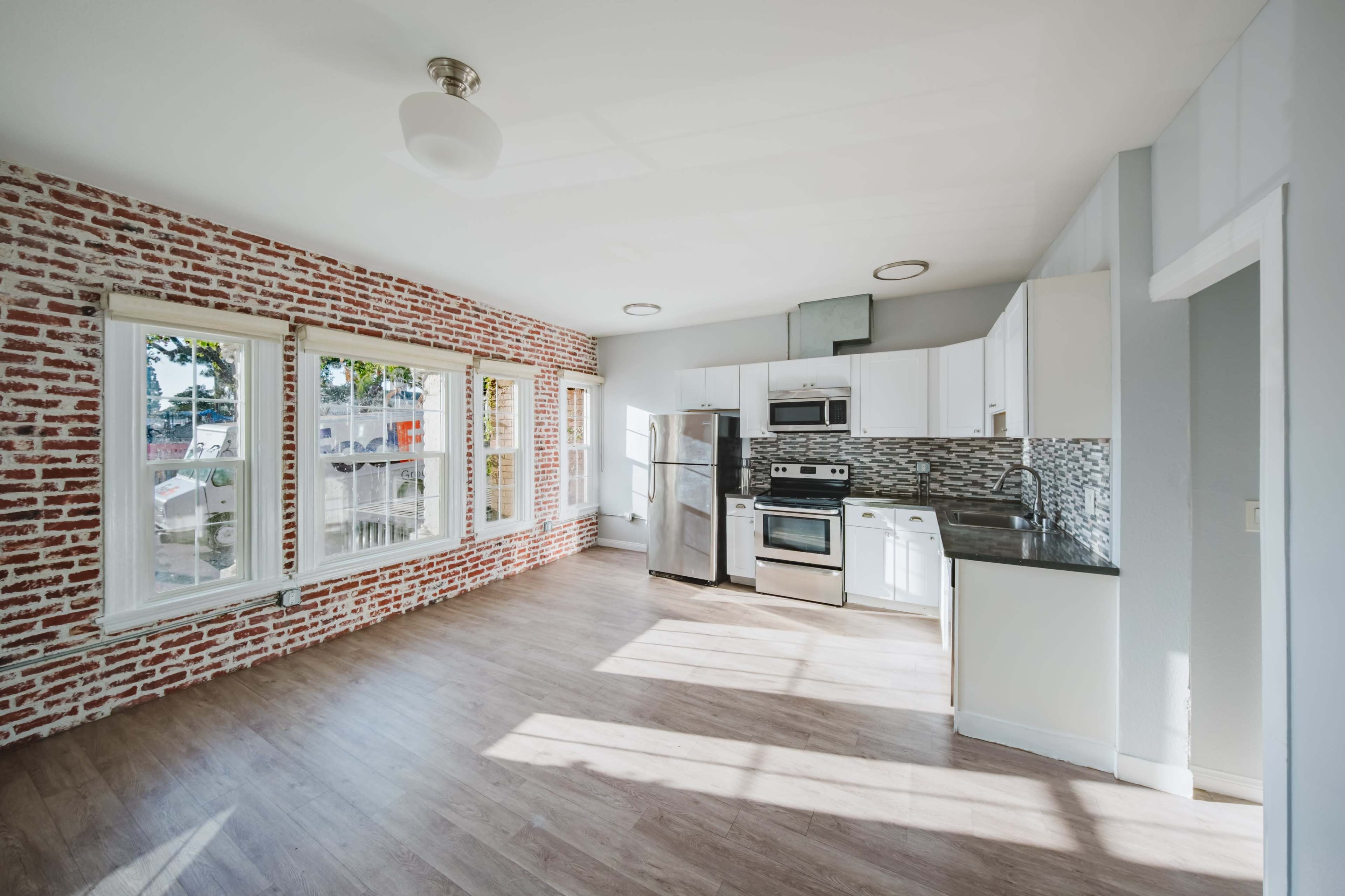 The image shows a modern kitchen with exposed brick walls, stainless steel appliances, and large windows allowing natural light to fill the space.
