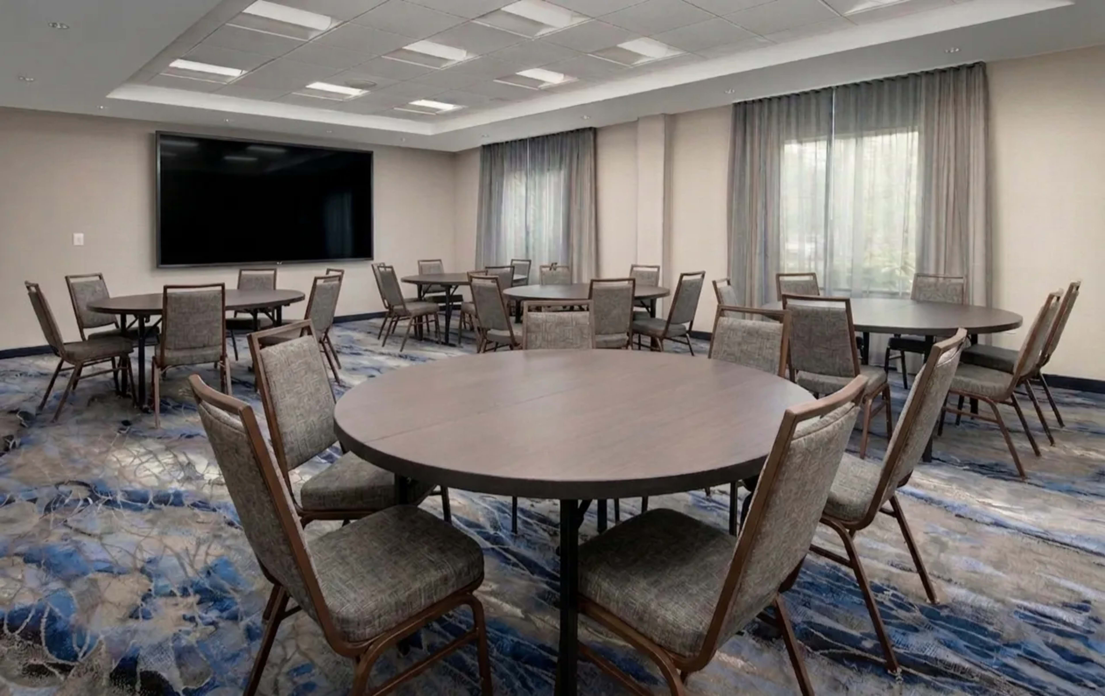 The image displays a conference room with multiple round tables arranged and surrounded by chairs, with large windows allowing natural light and a screen mounted on the wall.