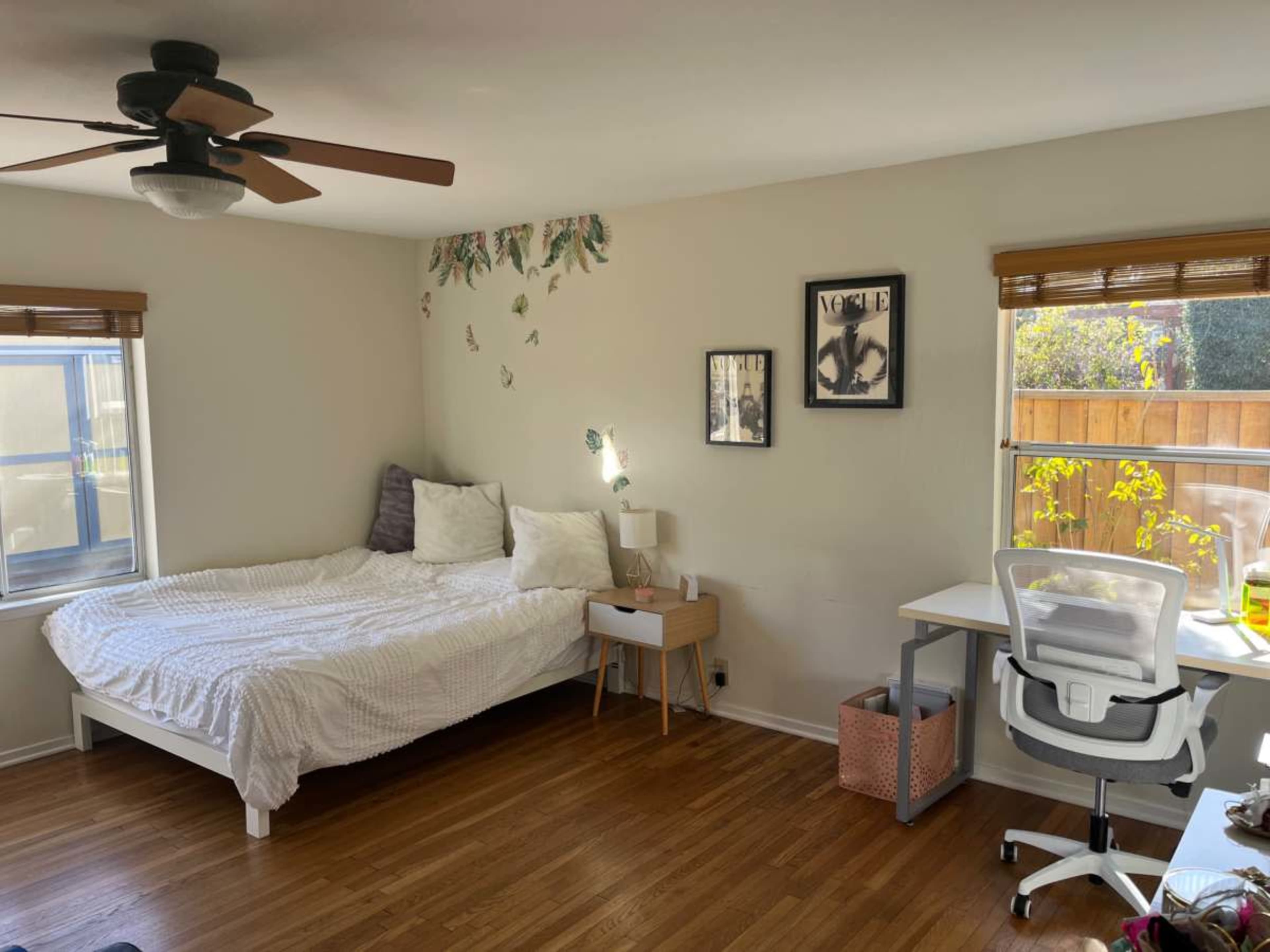 The image shows a bright, minimalistic bedroom featuring a bed, a desk with a chair, and natural light streaming through the windows.