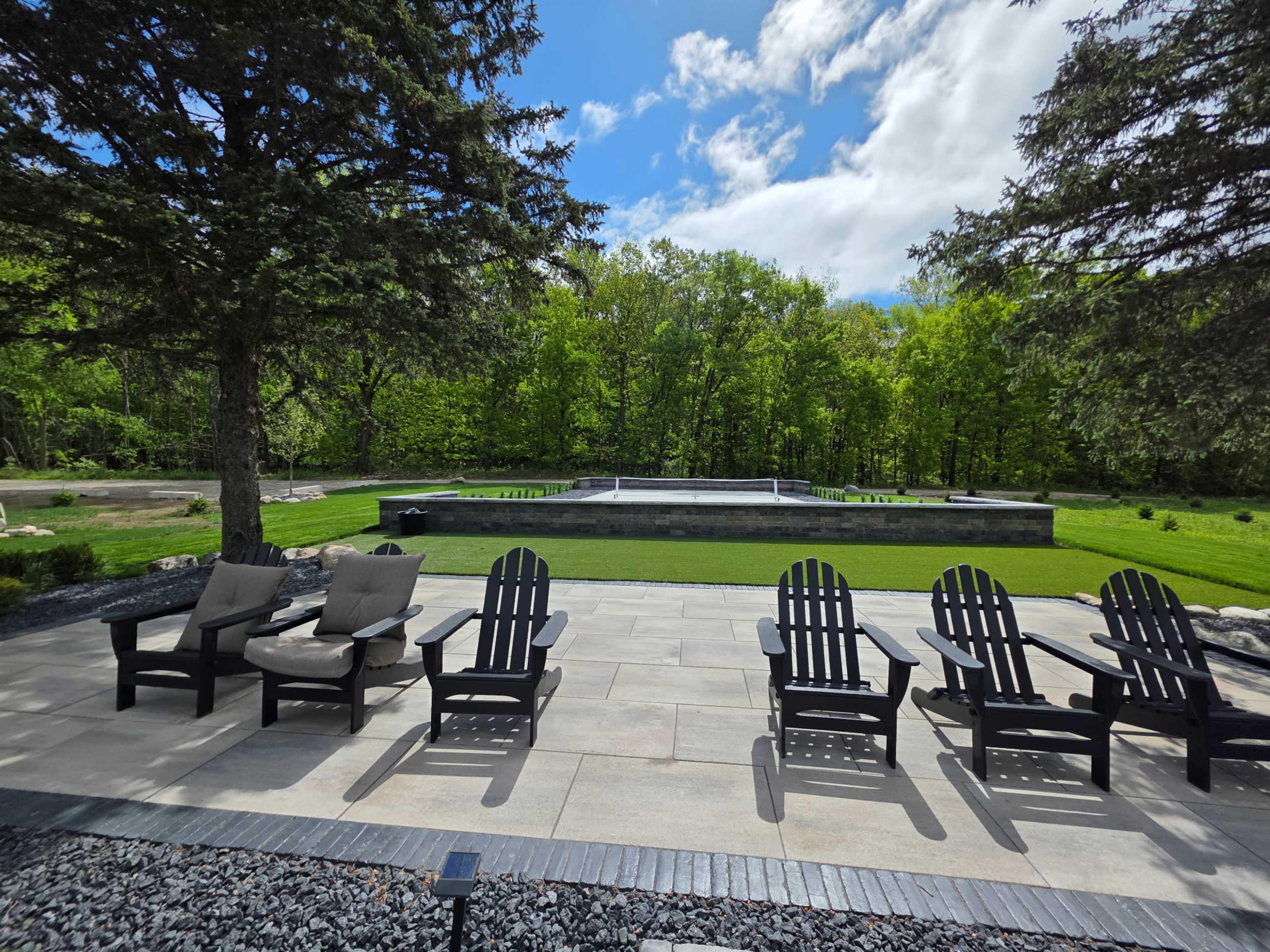 A patio with black Adirondack chairs faces a rectangular water feature surrounded by green trees and grass.