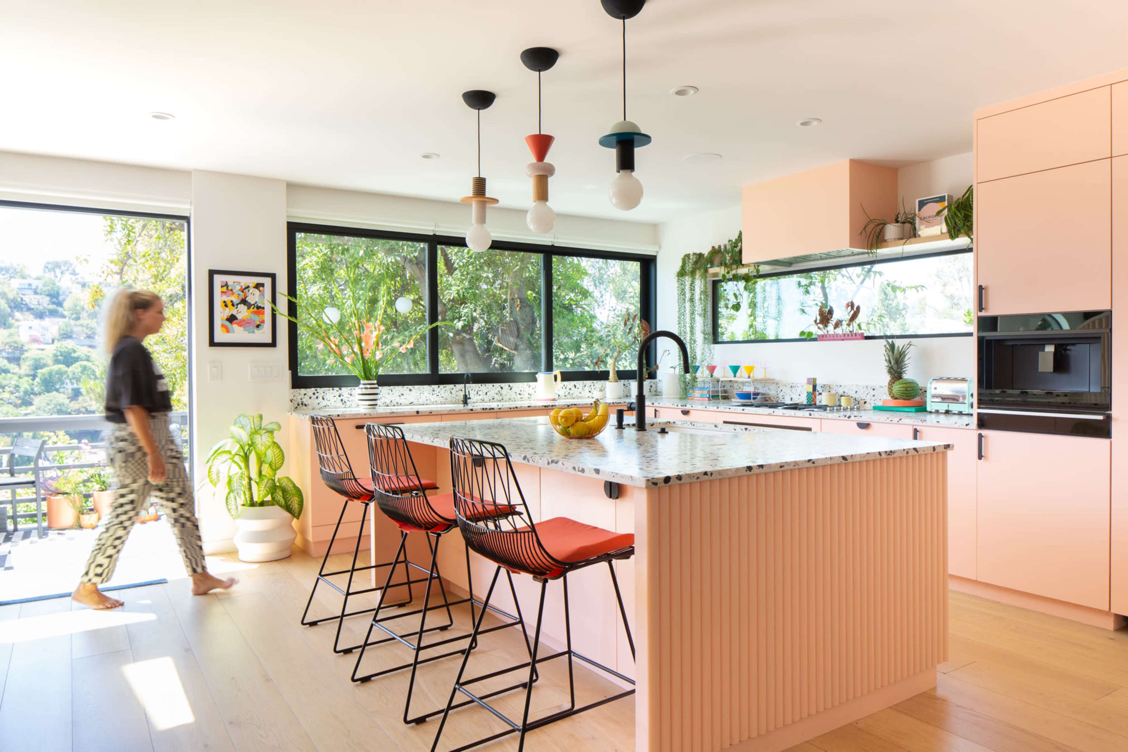 A modern kitchen features a large island with barstools, abundant natural light, and views of greenery through floor-to-ceiling windows.