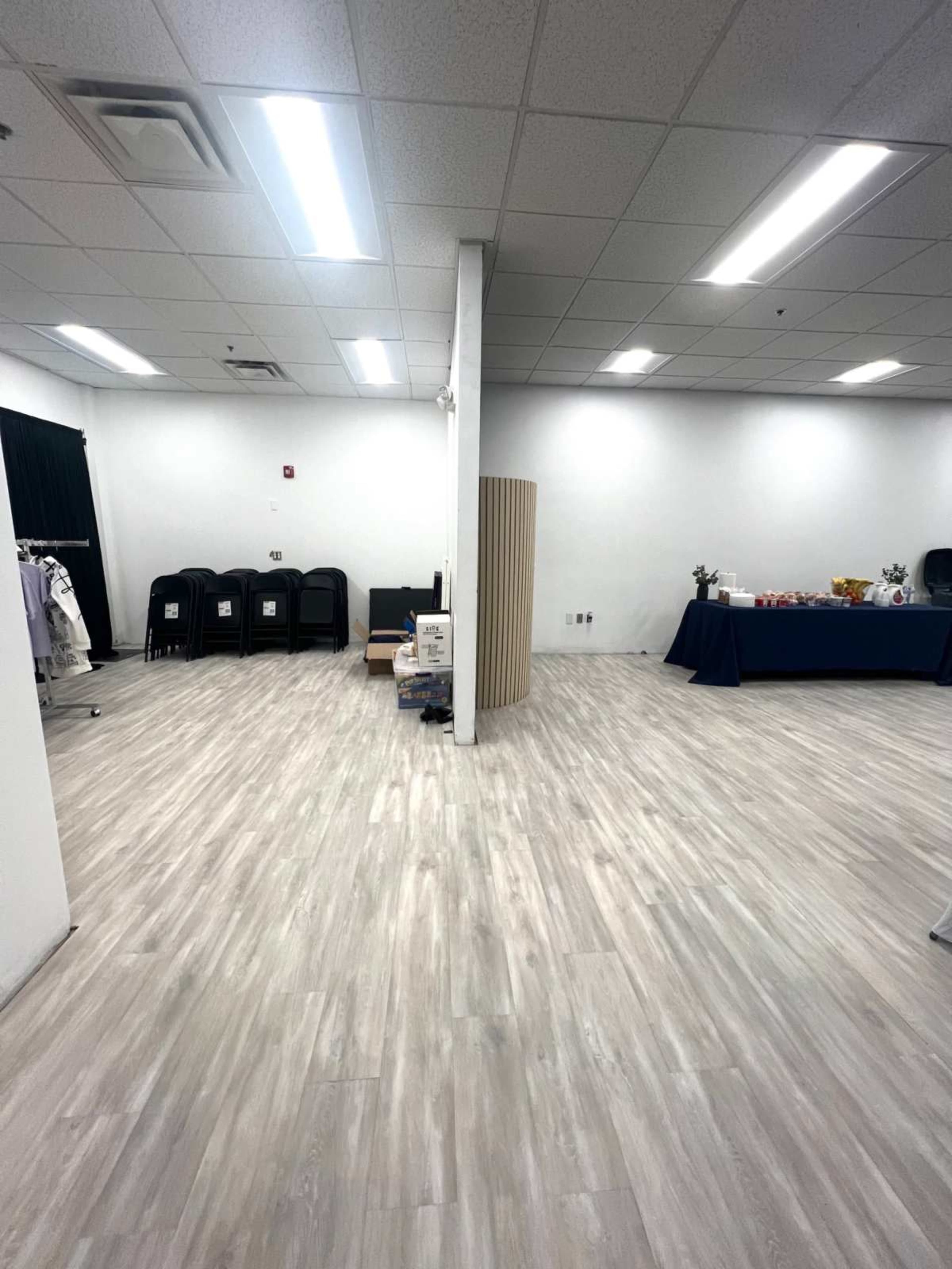 The image shows a spacious, empty room with light wood flooring and sectioned areas for seating and a table setup.