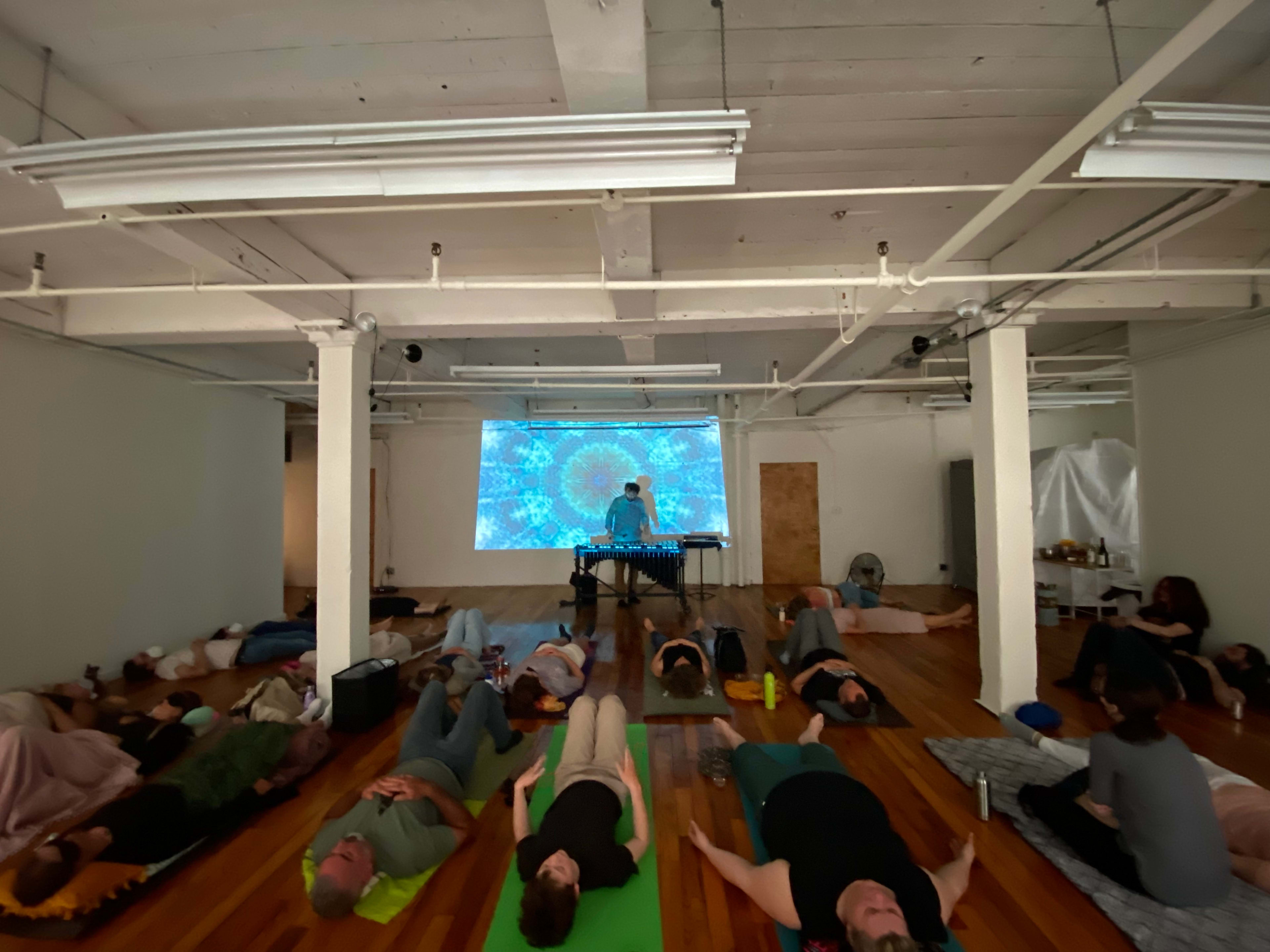 A group of people lies on yoga mats in a room with a large projection on the wall, featuring abstract visuals, while an instructor plays a musical instrument at the front.