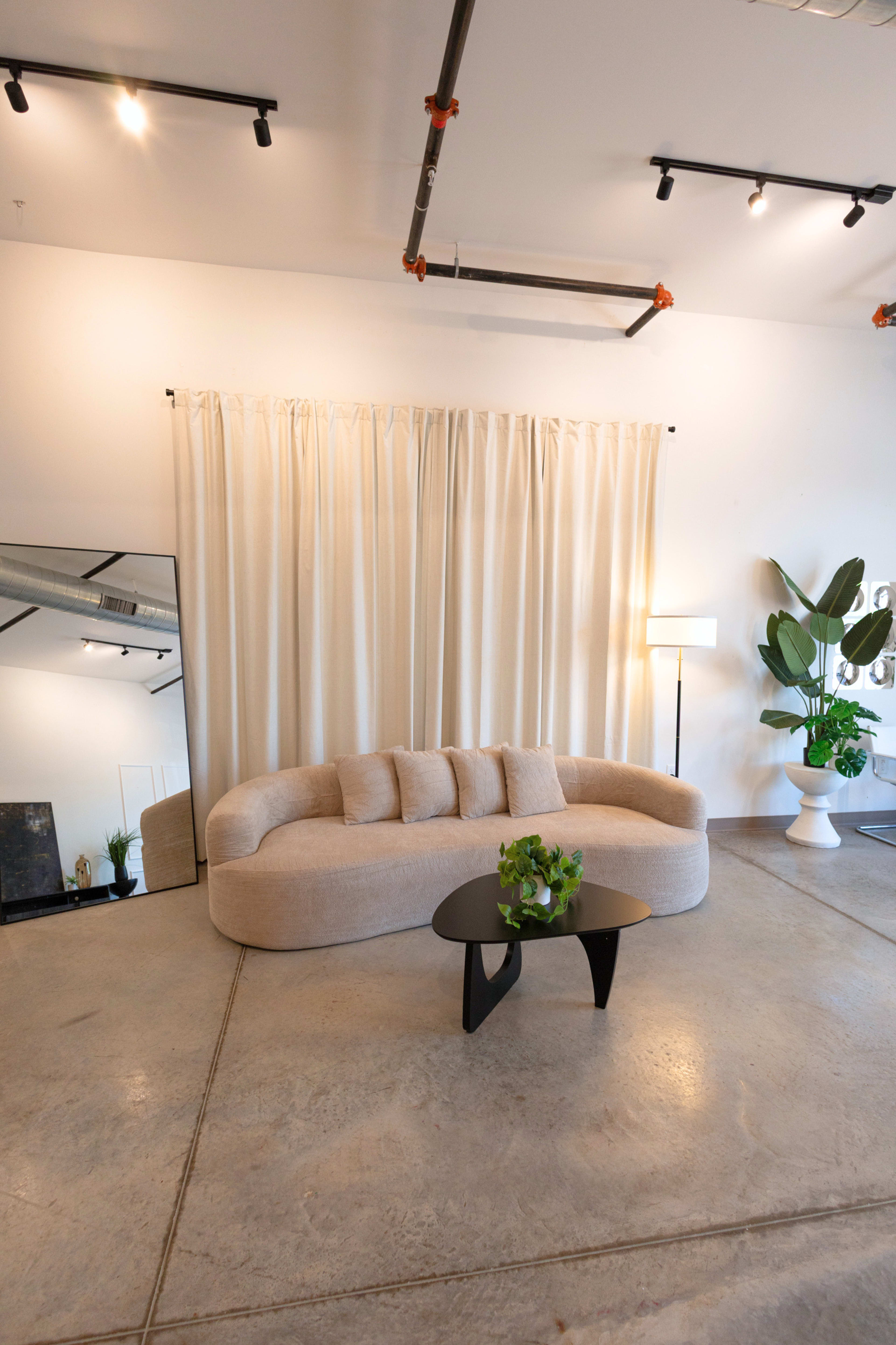 A curved beige sofa is positioned in front of a large mirror, with white curtains and a potted plant nearby in a contemporary interior space.