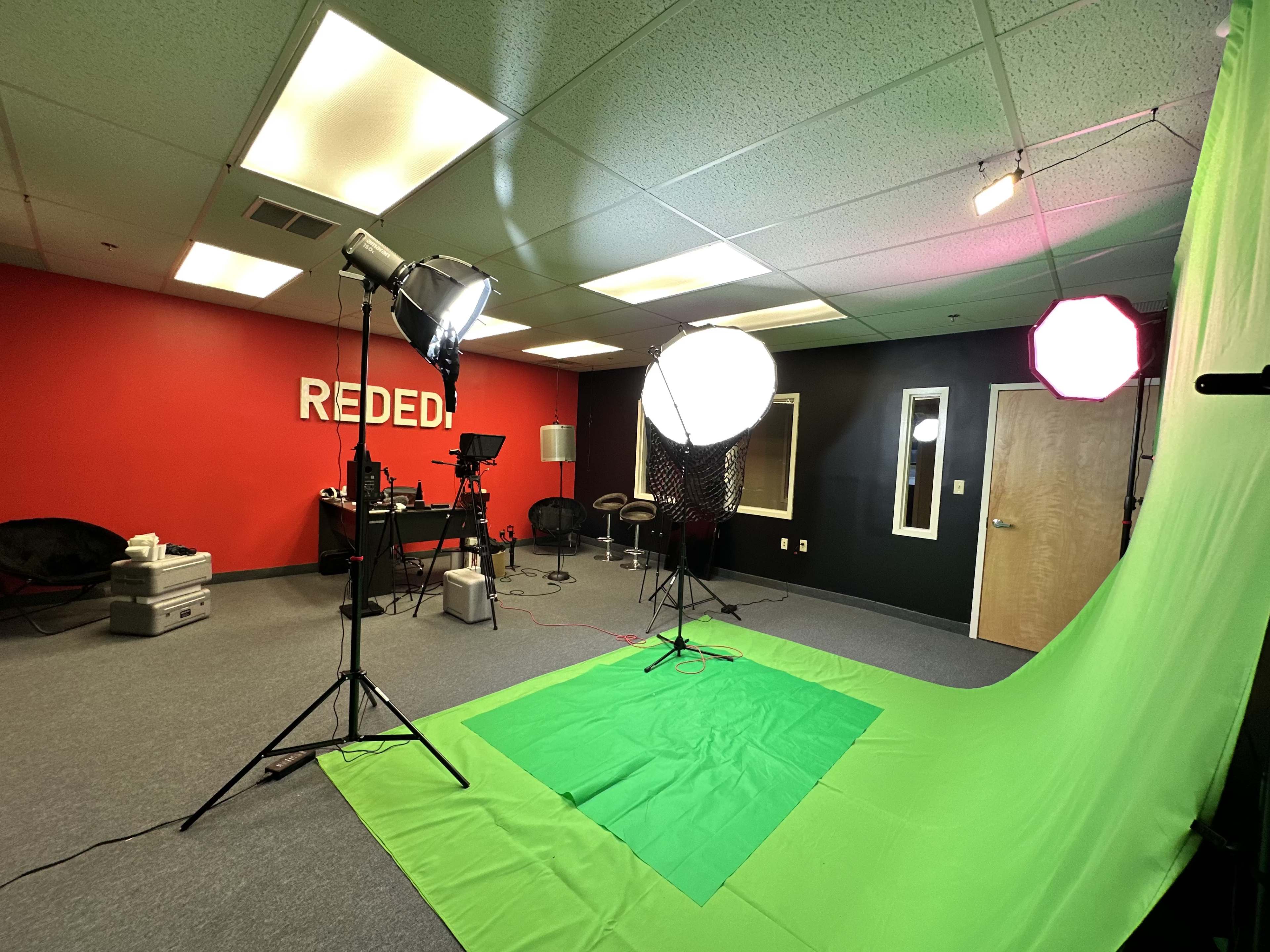 The image shows a studio setup with a green backdrop, lighting equipment, and a desk area against a red wall.