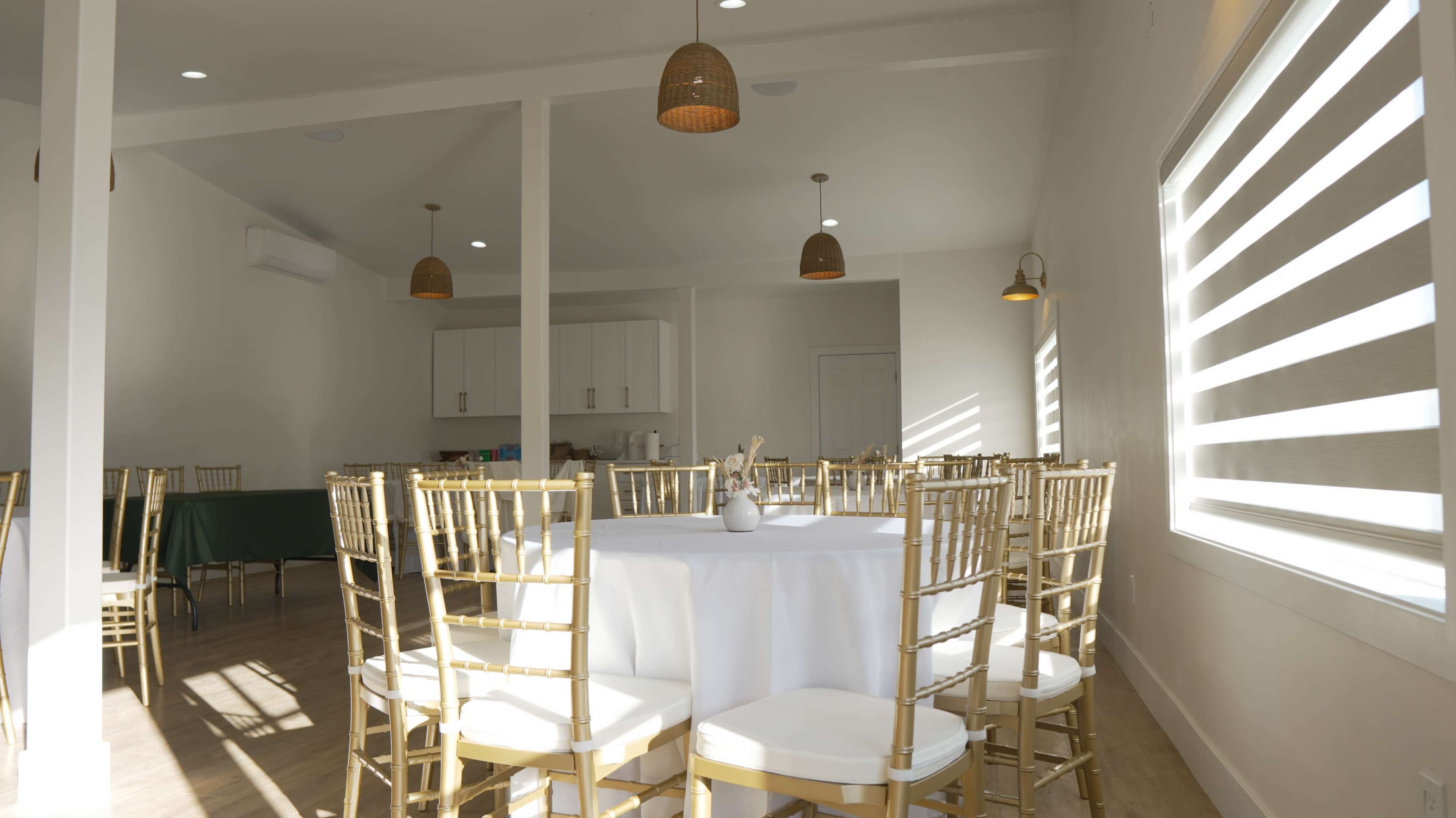 A bright, modern event space features a round table set with a white tablecloth and surrounded by gold chairs, illuminated by pendant lighting.
