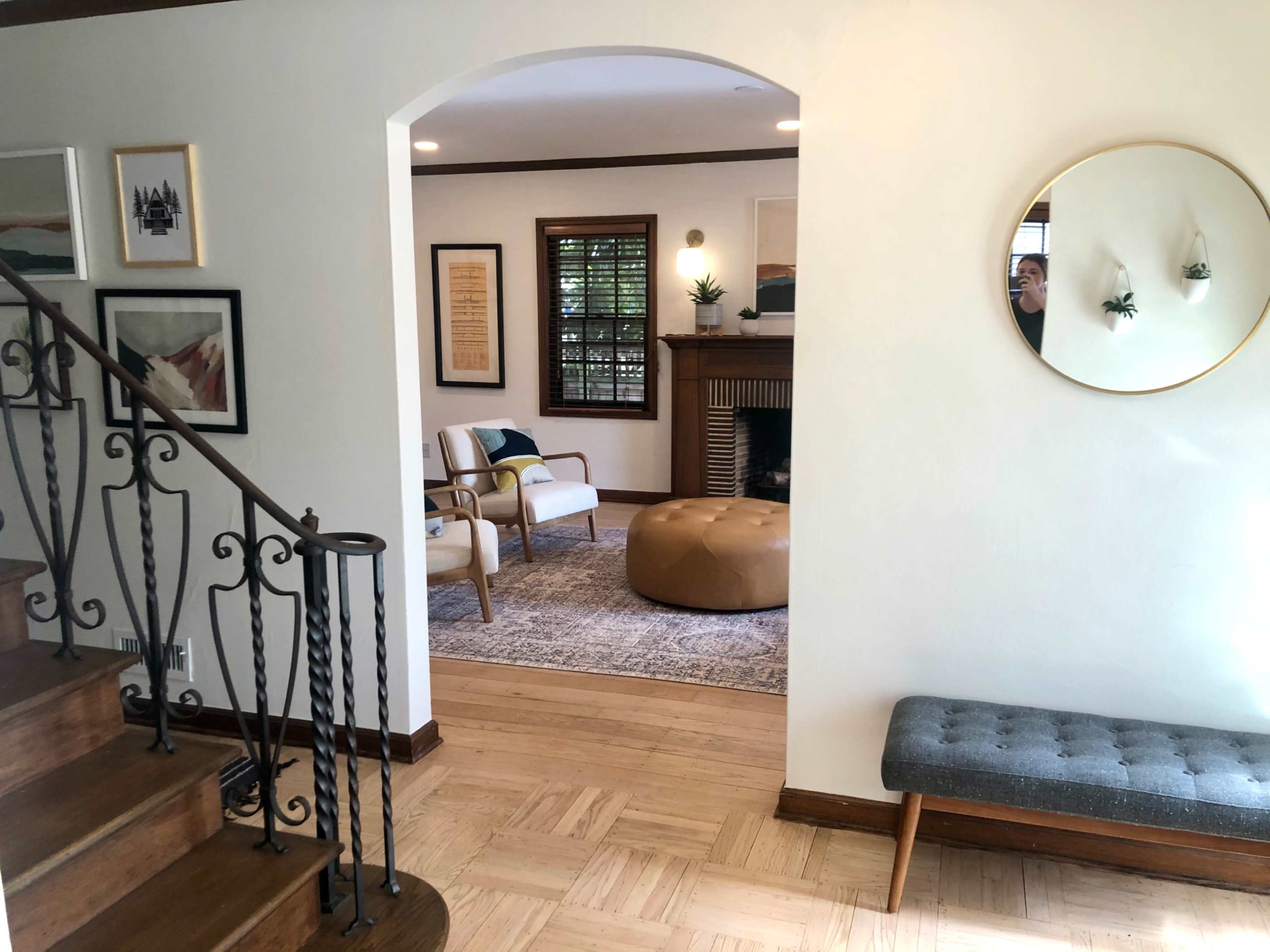A living space featuring a staircase, a round mirror, and a seating area with two chairs, a large ottoman, and artwork on the walls.