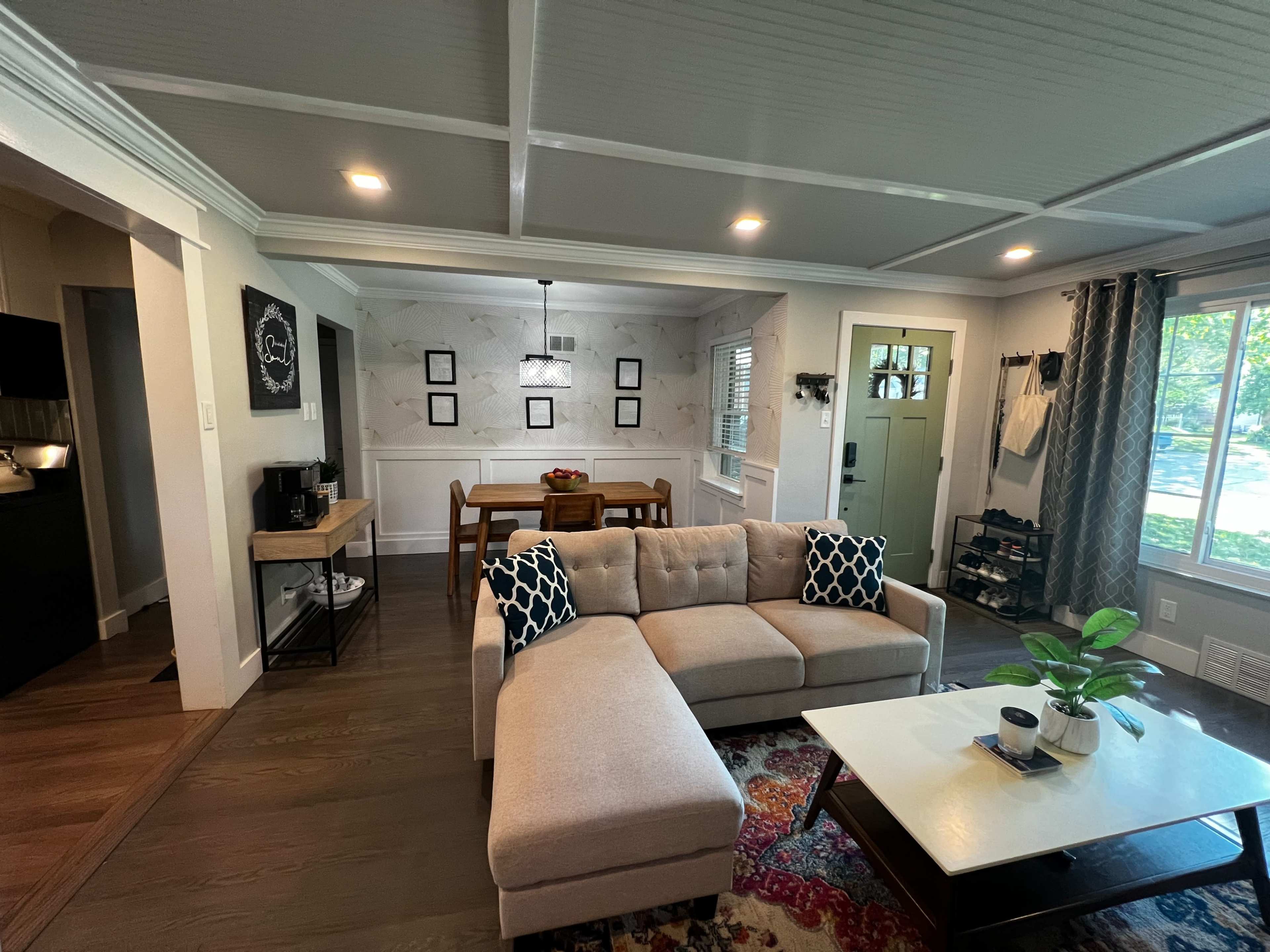 A spacious living room with a sectional sofa, a coffee table, and a dining area adjacent to a kitchen, illuminated by natural light from nearby windows.