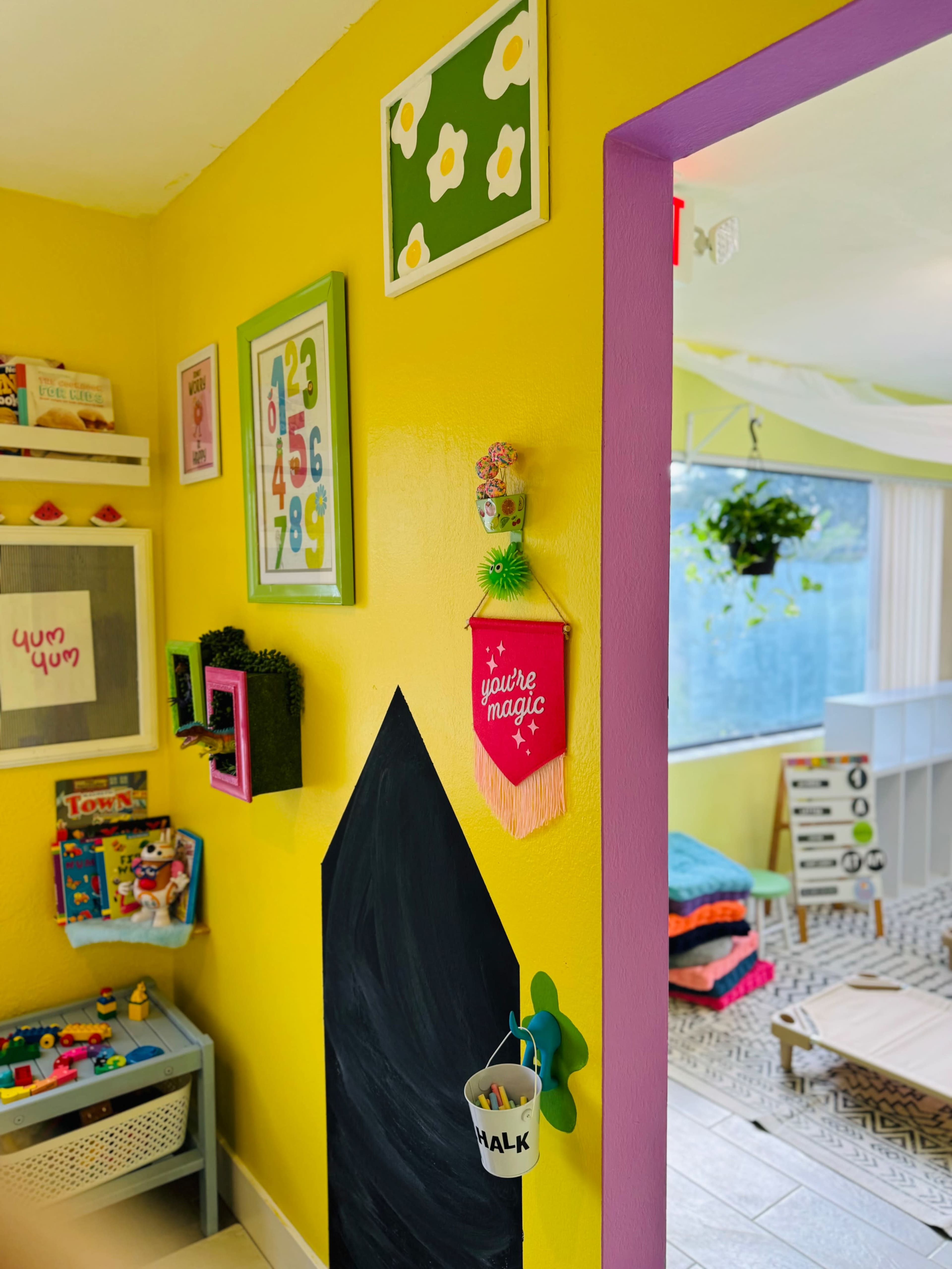 The image shows a brightly colored kids' playroom with a yellow wall, framed artwork, and a pink-trimmed entrance to another area.