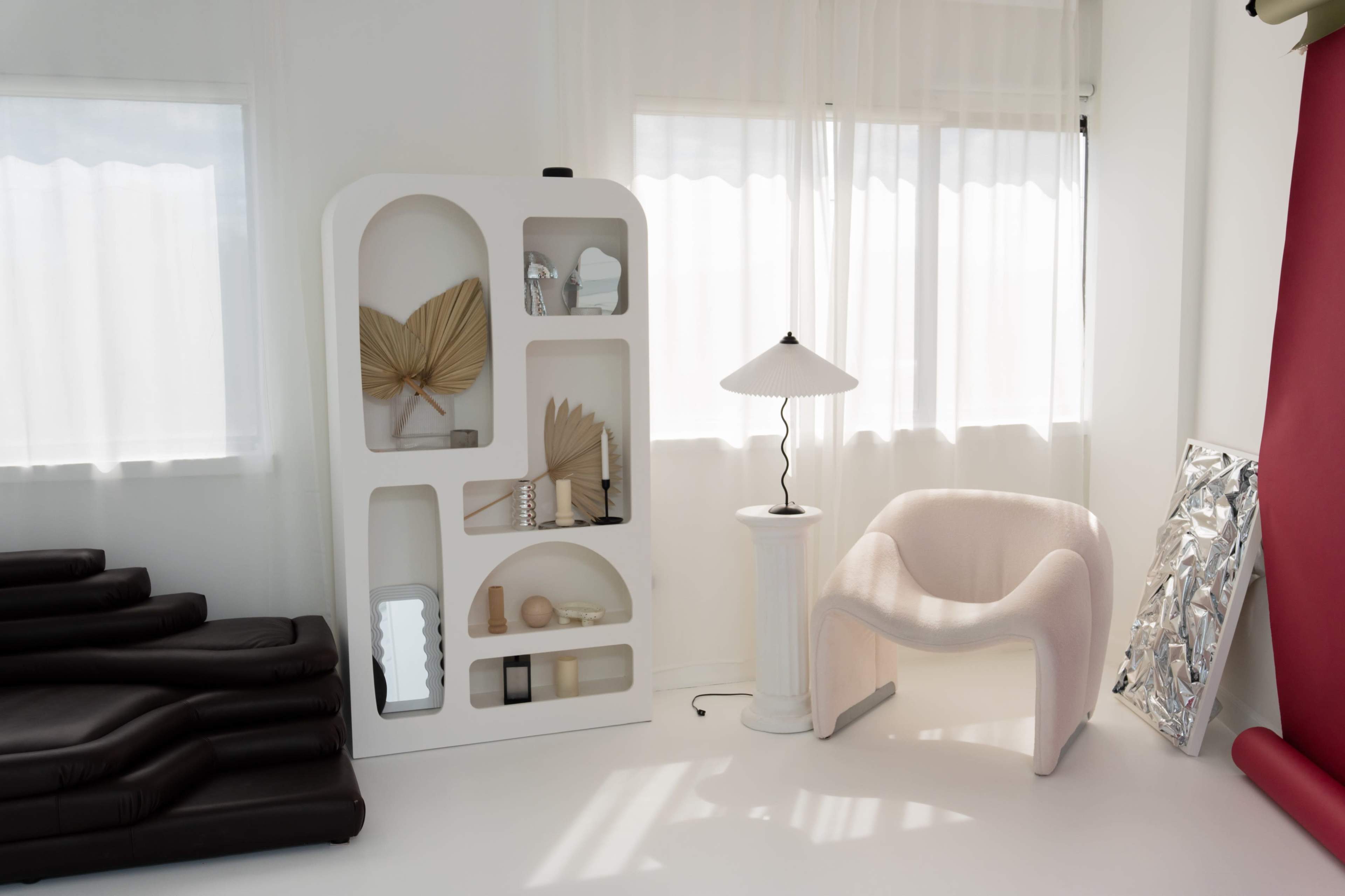 The image shows a minimalist interior with a white shelving unit, a lamp on a pedestal, a curved armchair, and a reflection of a shiny surface, all set against light-colored walls and large windows.