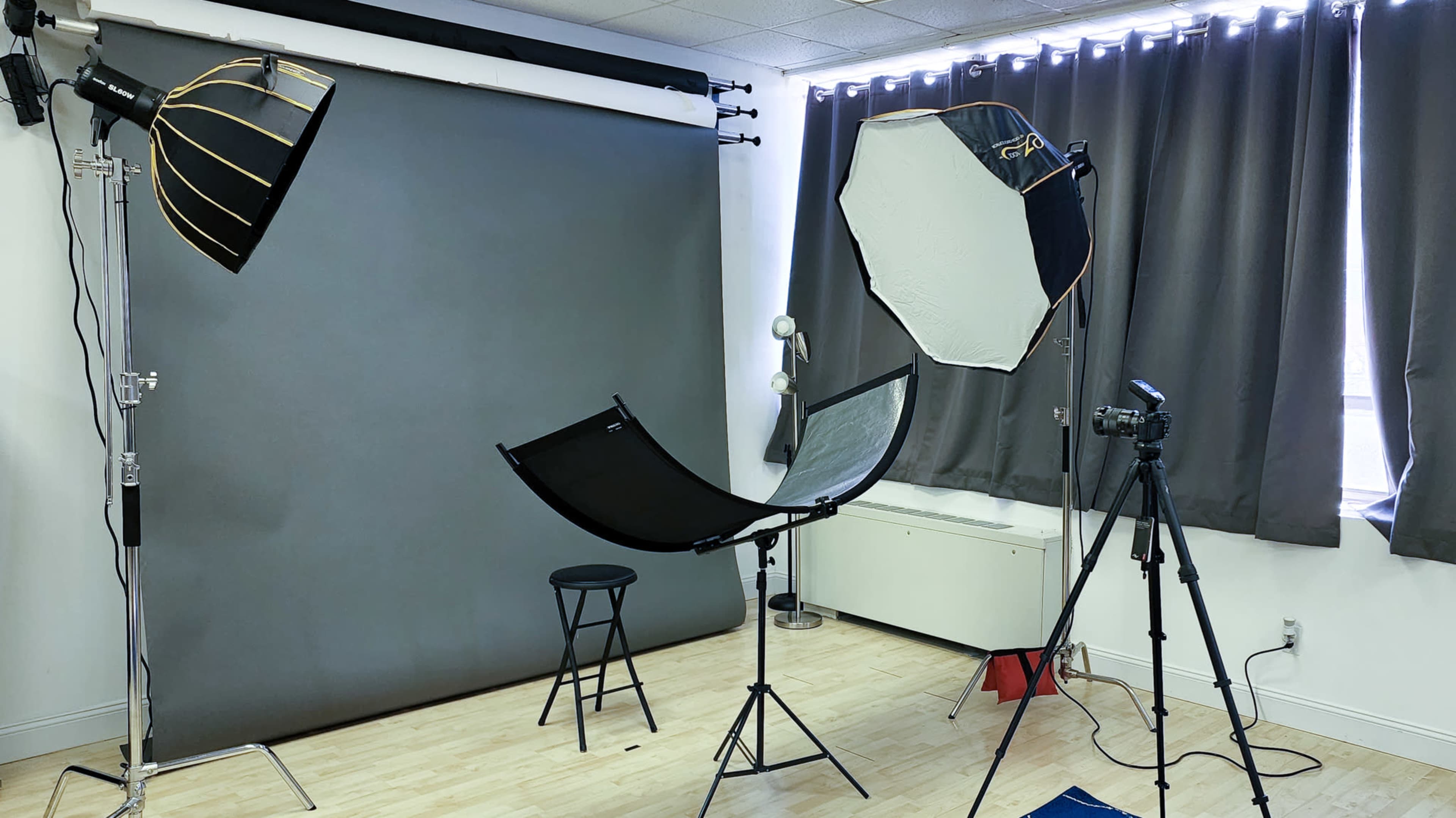 A photography studio setup with lighting equipment, a backdrop, a reflector, a chair, and a camera on a tripod.