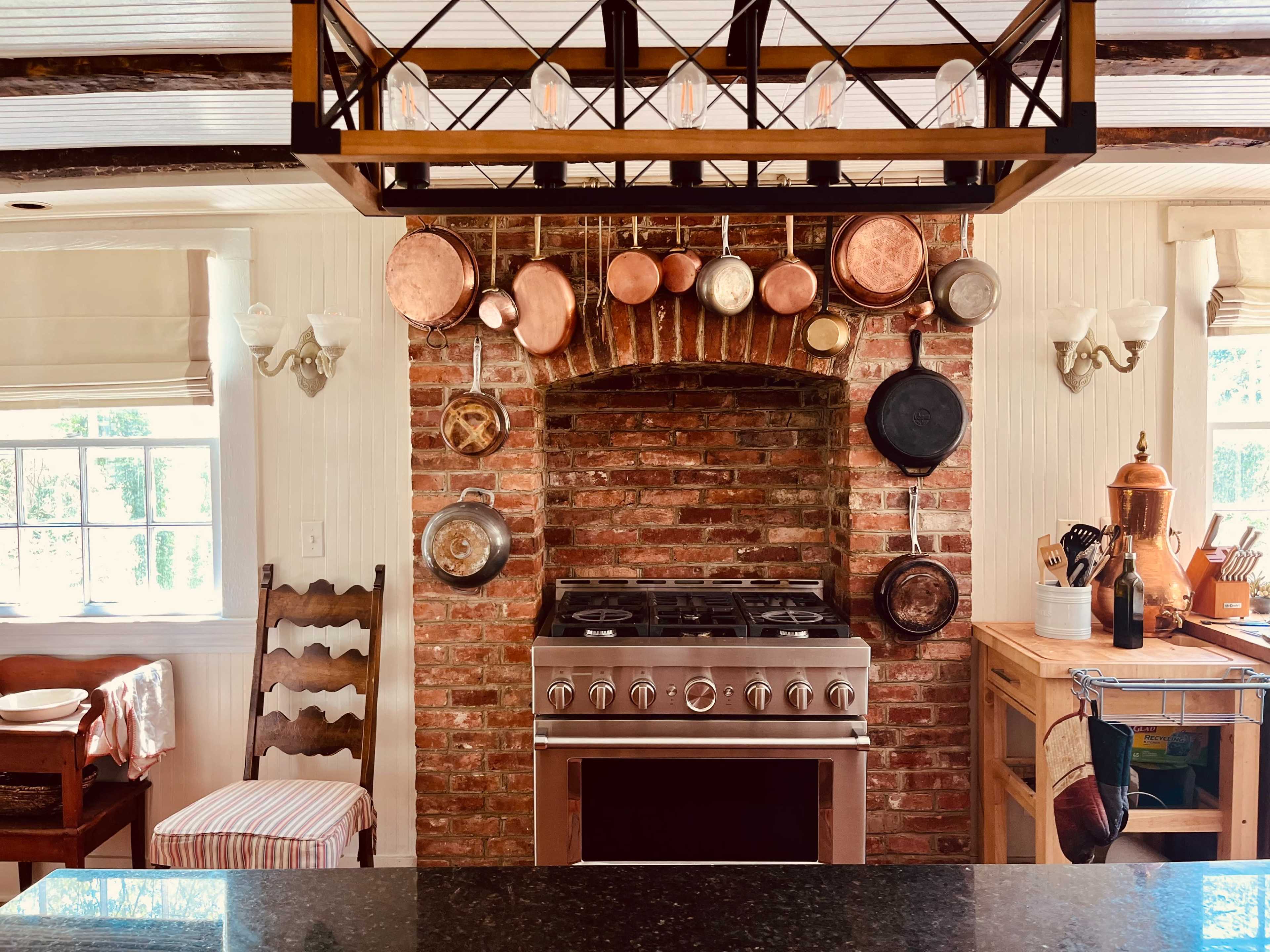 A rustic kitchen with a brick oven, hanging copper cookware, and a view of wooden furniture.