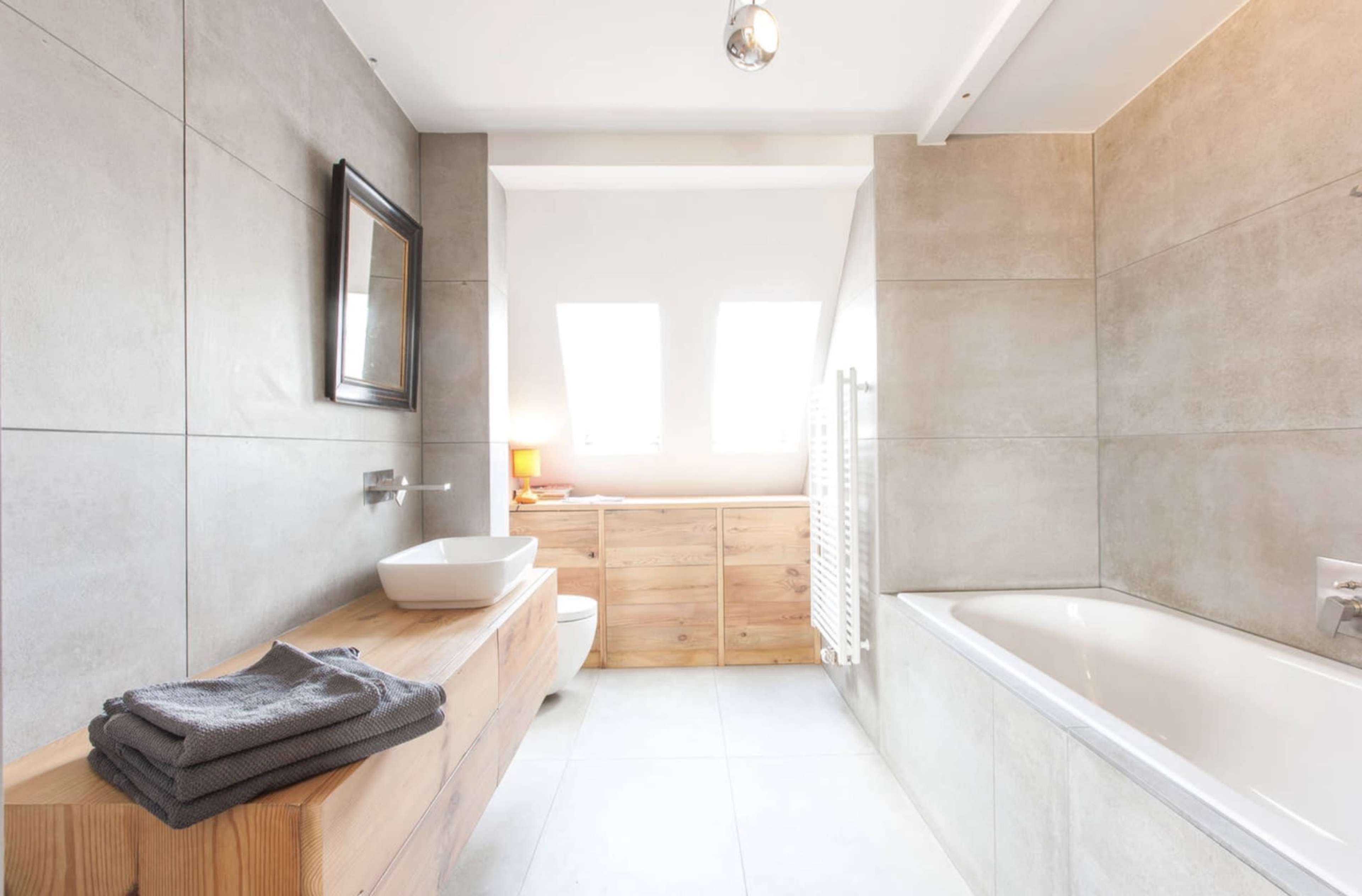 The image shows a modern bathroom with gray tiled walls, a wooden vanity, a white bathtub, and a skylight allowing natural light in.