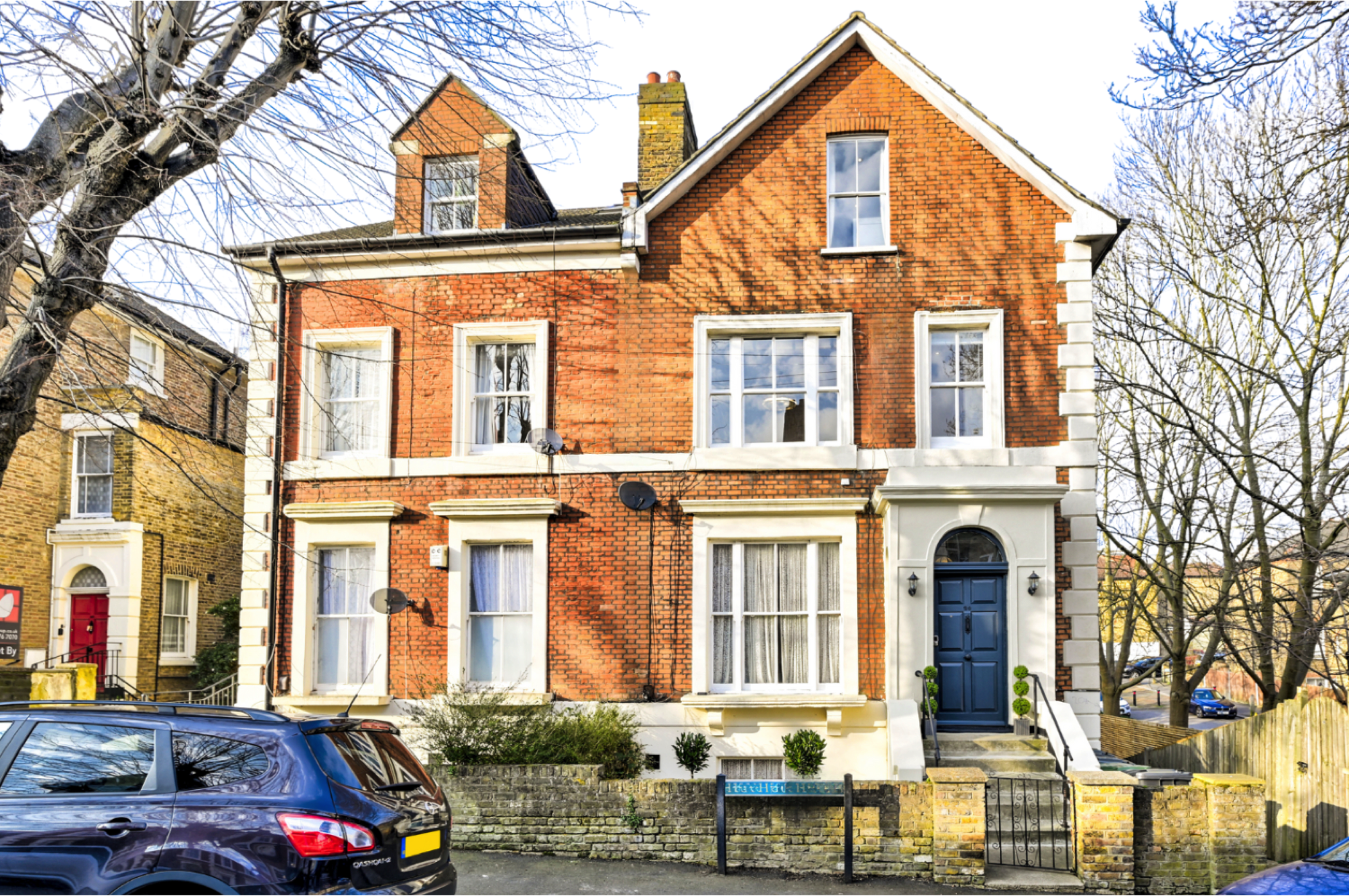 A three-story brick townhouse featuring large windows and a blue front door, situated among bare trees and parked cars.