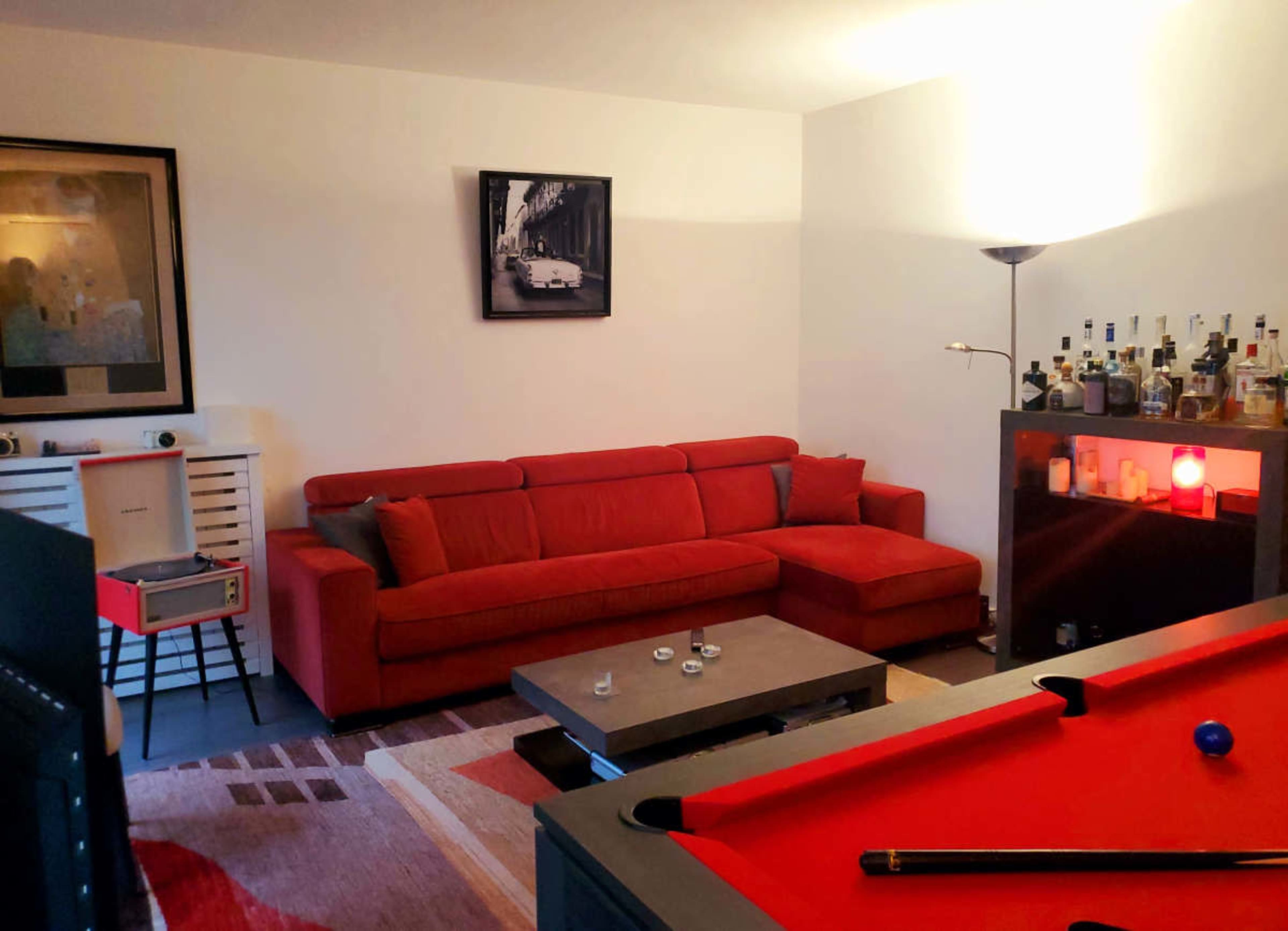 The image shows a cozy living room with a red sofa, a coffee table, and a pool table, complemented by a soft light and shelves displaying various bottles.