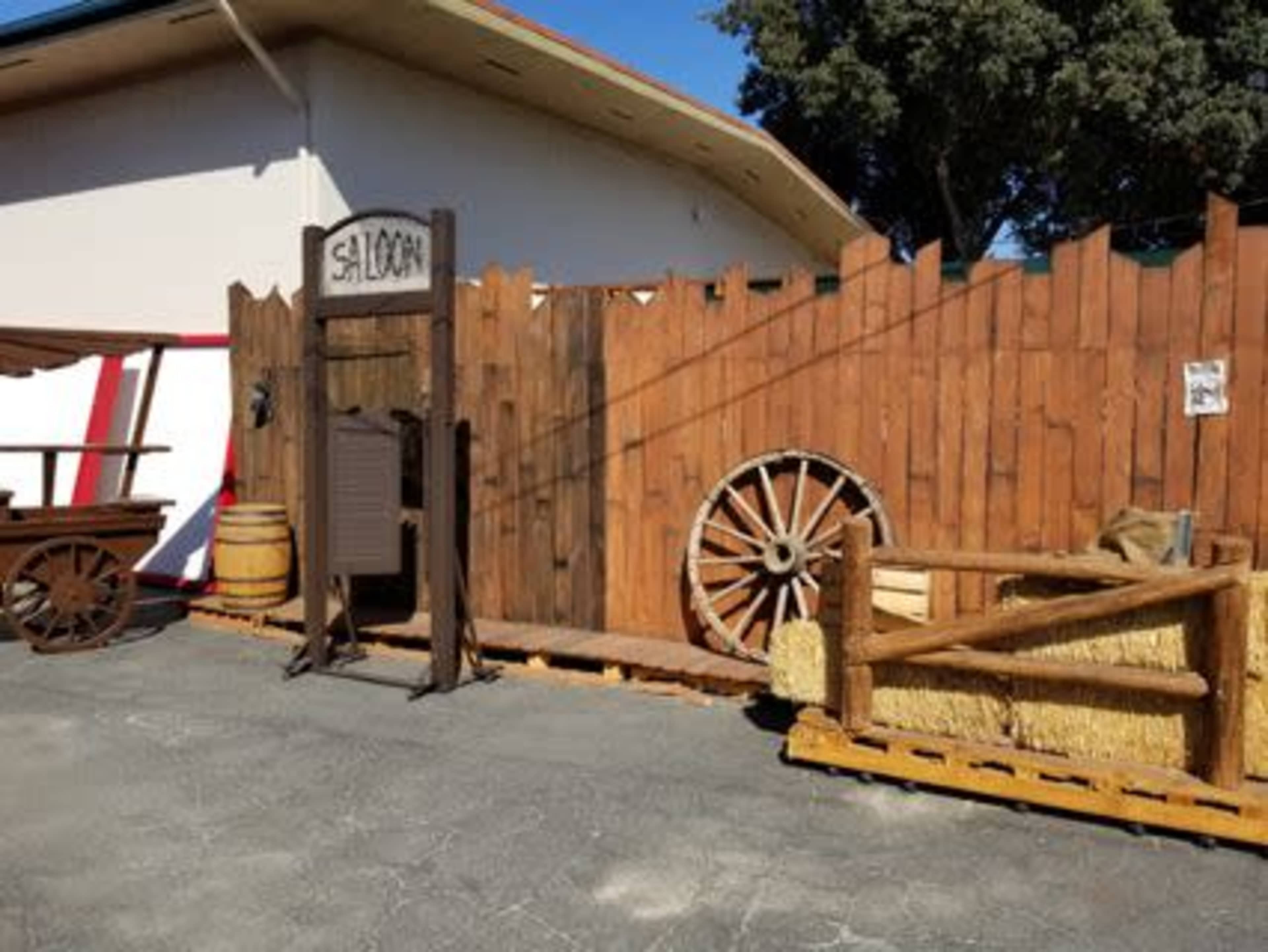A western-style set with a wooden saloon sign, a wagon wheel, barrels, and straw bales against a rustic wooden fence.