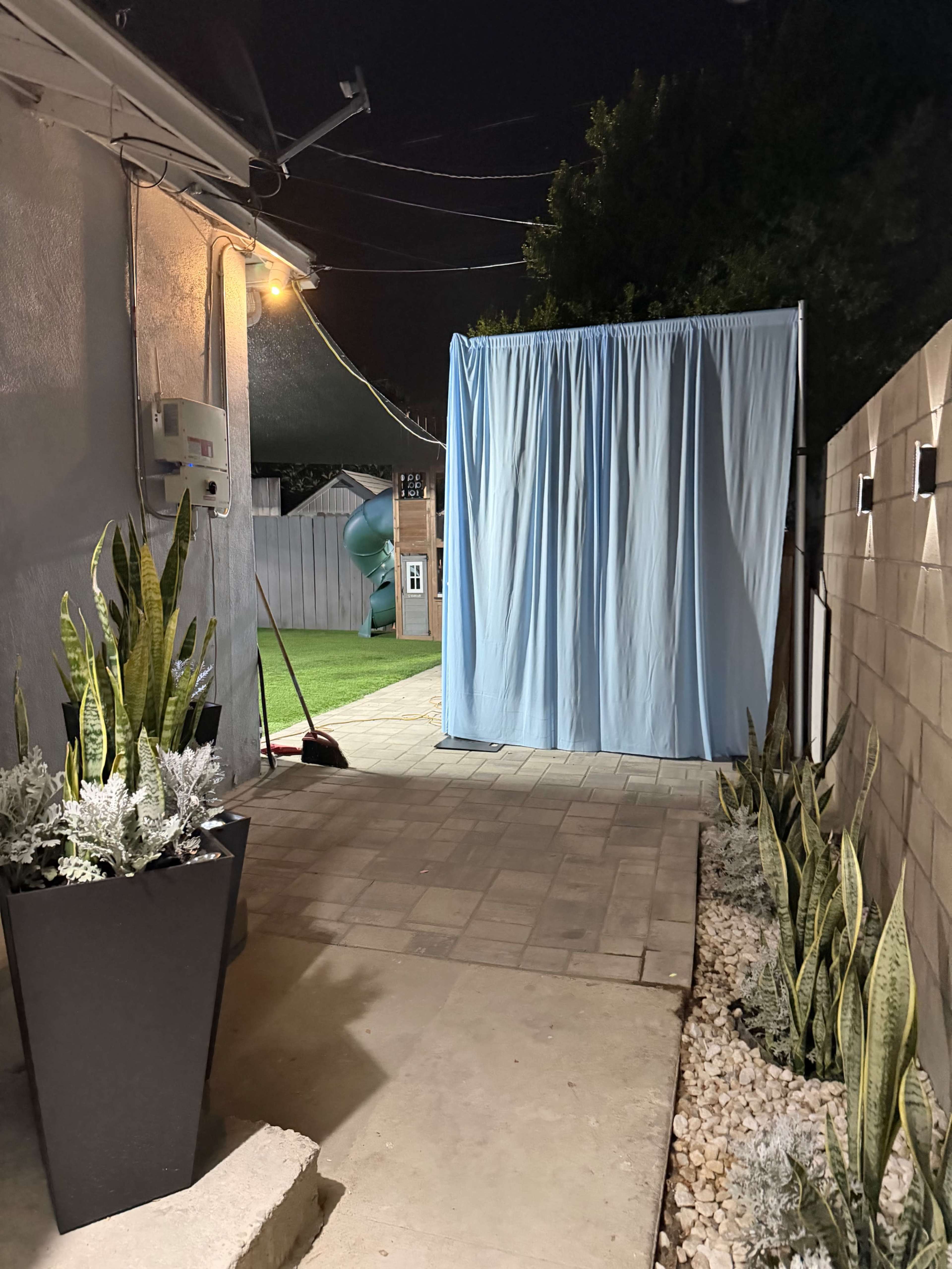 A pathway leads from a house to a green lawn, where a blue curtain drapes over a structure, with planters on either side.